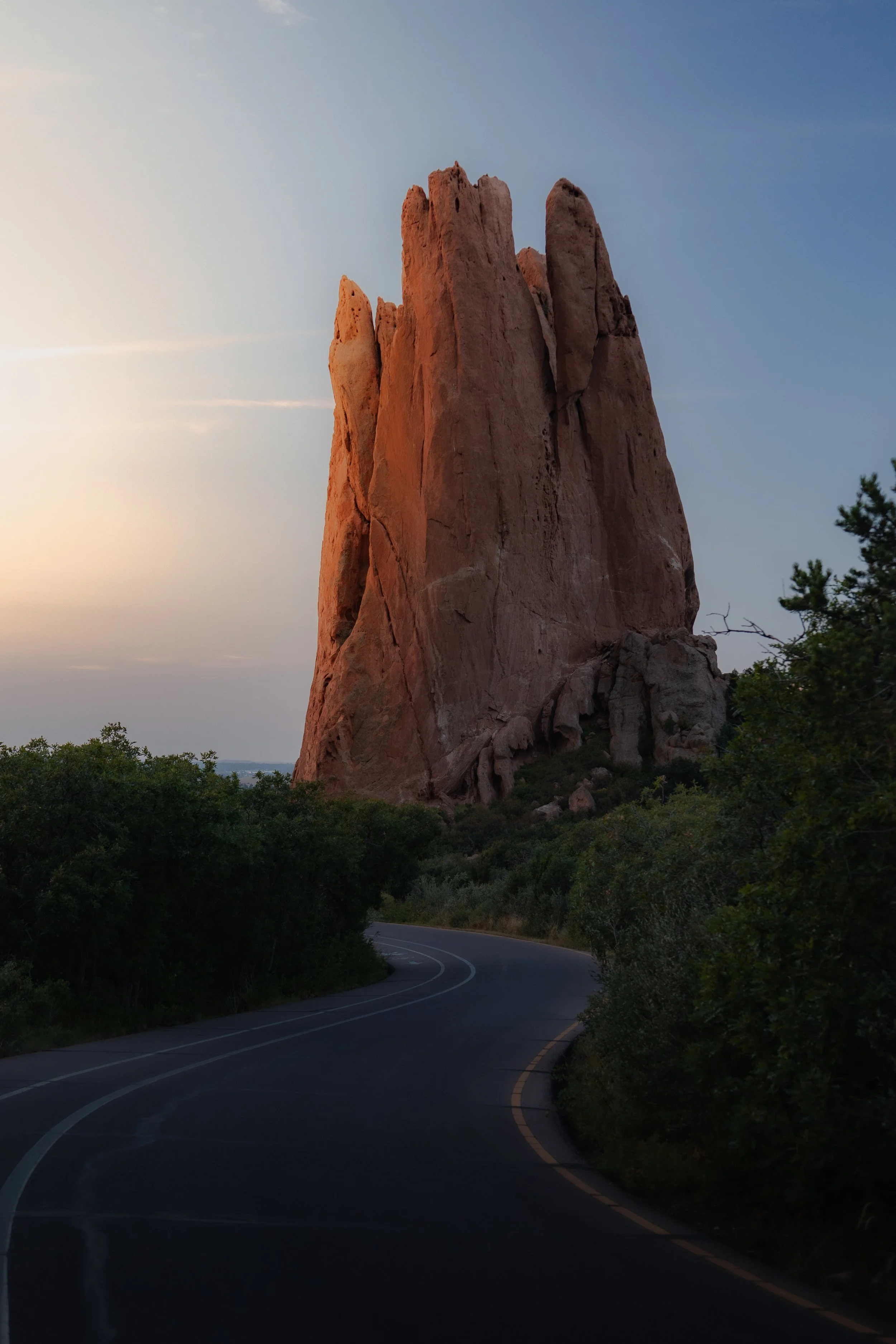 A large red rock formation towering over a winding road with green trees on either side, under a clear sky at dusk.