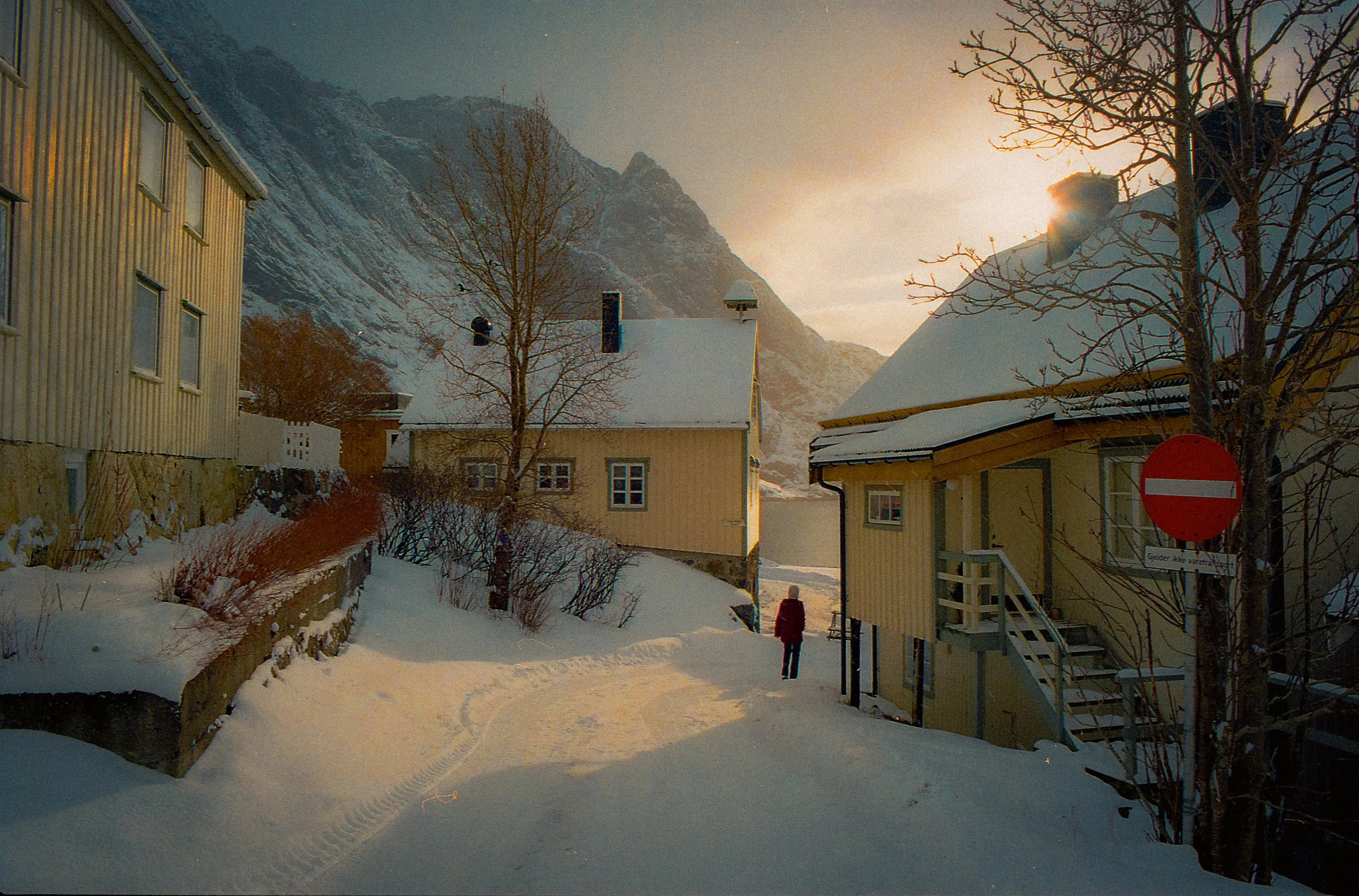 Snow-covered residential area with yellow and white wooden houses, leafless trees, and a mountain backdrop at sunset. A person in red walking on the snow path.