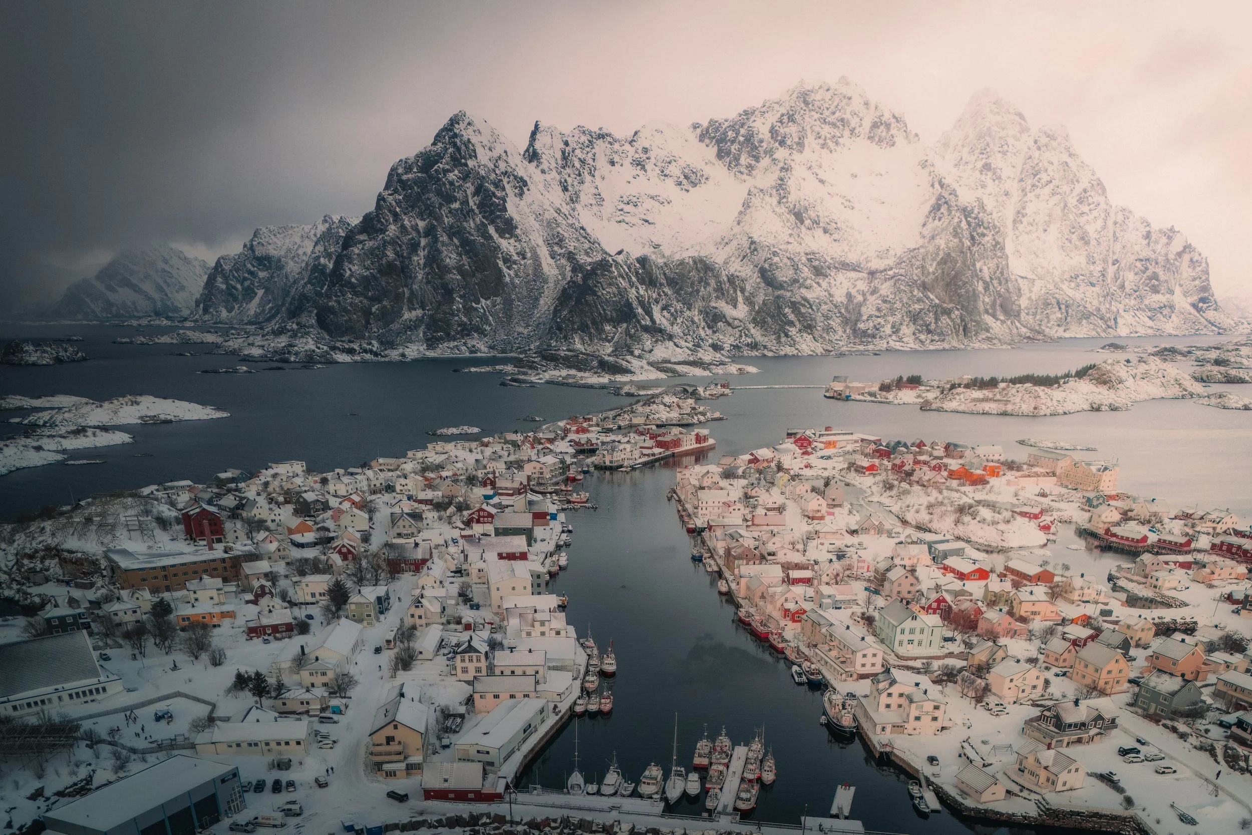 A snow-covered coastal village with colorful houses and boats, set against a backdrop of tall, rugged, snow-capped mountains.