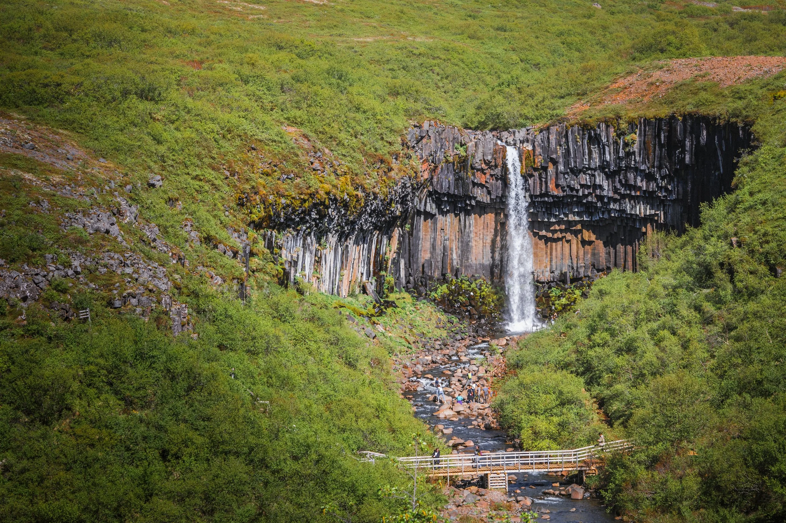Waterfall flowing over basalt columns in lush green landscape with a wooden bridge and people along the stream.