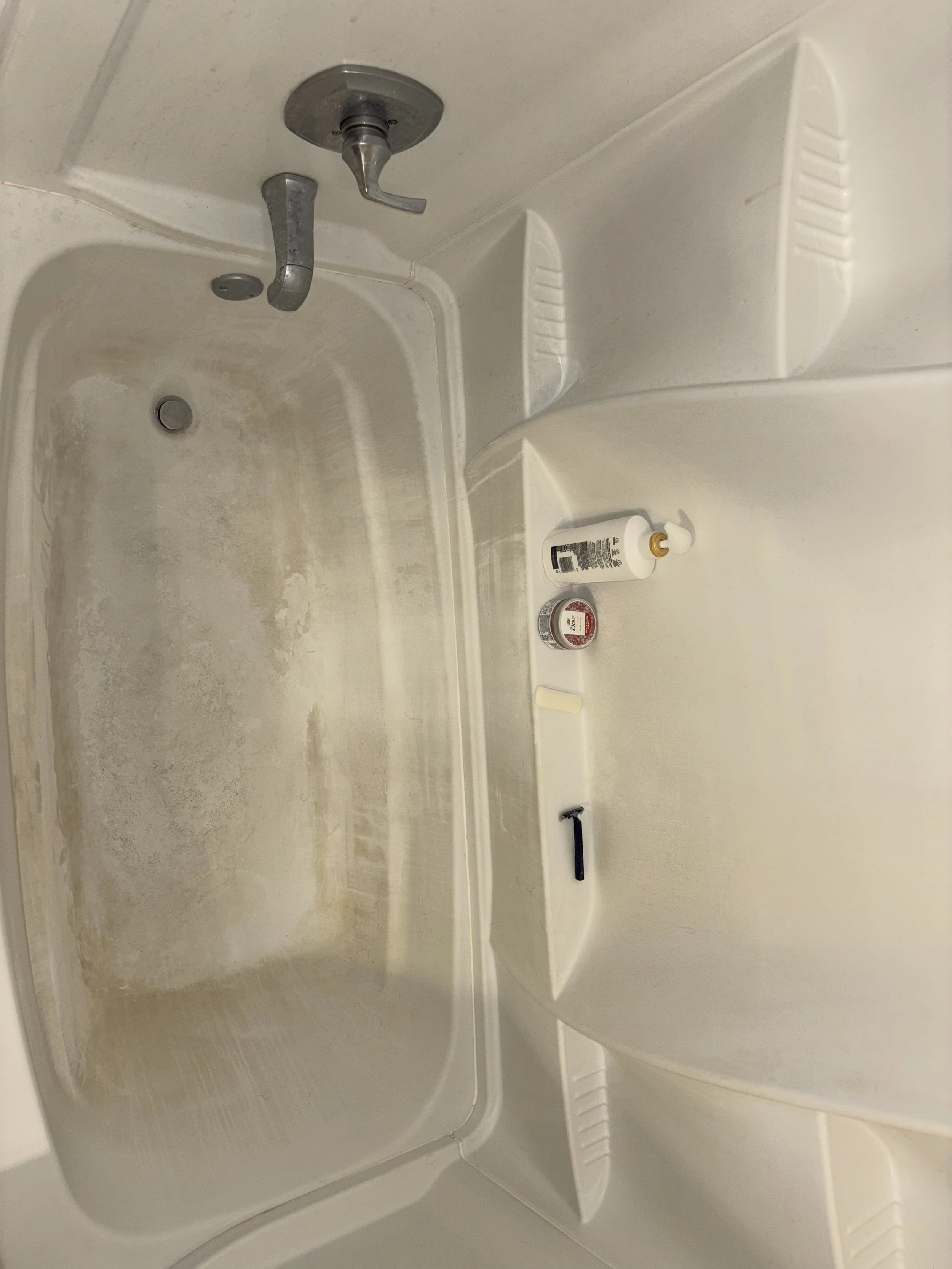 Inside a bathtub with some dirt and grime, with a faucet, shower handle, and a few toiletries on the built-in ledge.