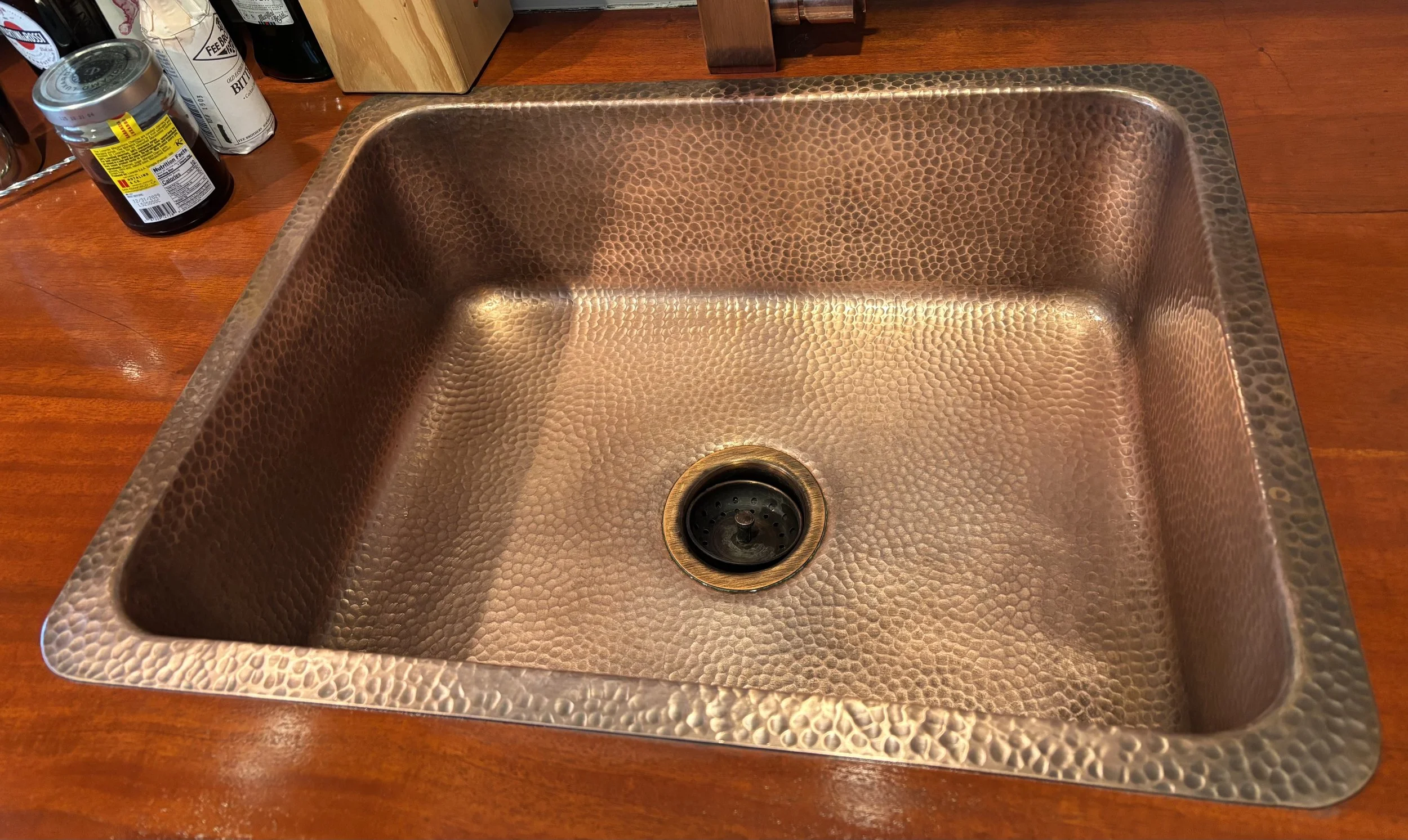 A hammered copper kitchen sink restored from hard buildup, installed on a wooden countertop near some jars and bottles.