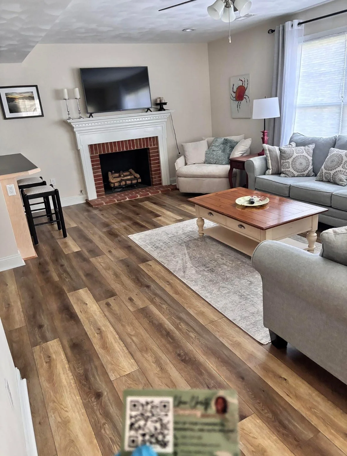 Living room with wood flooring, a white fireplace with a mounted flat-screen TV, a sectional sofa with decorative pillows, a coffee table, and a corner with a white armchair. Large window with white curtains, ceiling fan, and wall art of a red crab.