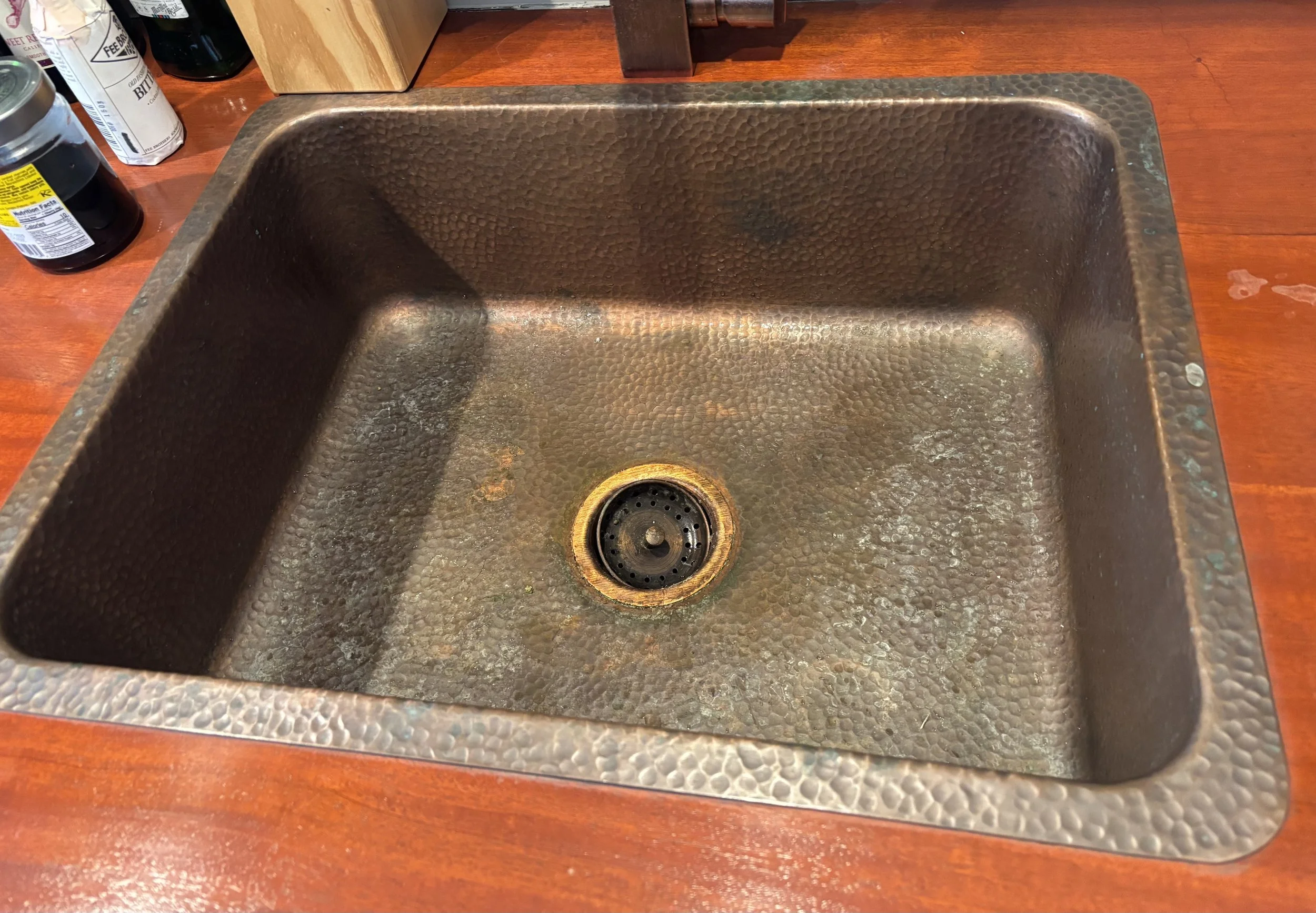 A large, hammered copper kitchen sink with a rusted look, installed in a wooden countertop.