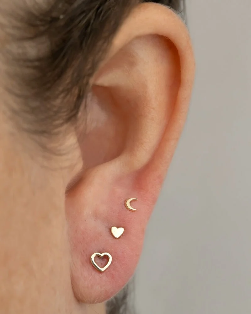 Close-up of an ear with three gold earrings: a crescent moon, a small heart, and a larger heart outline.