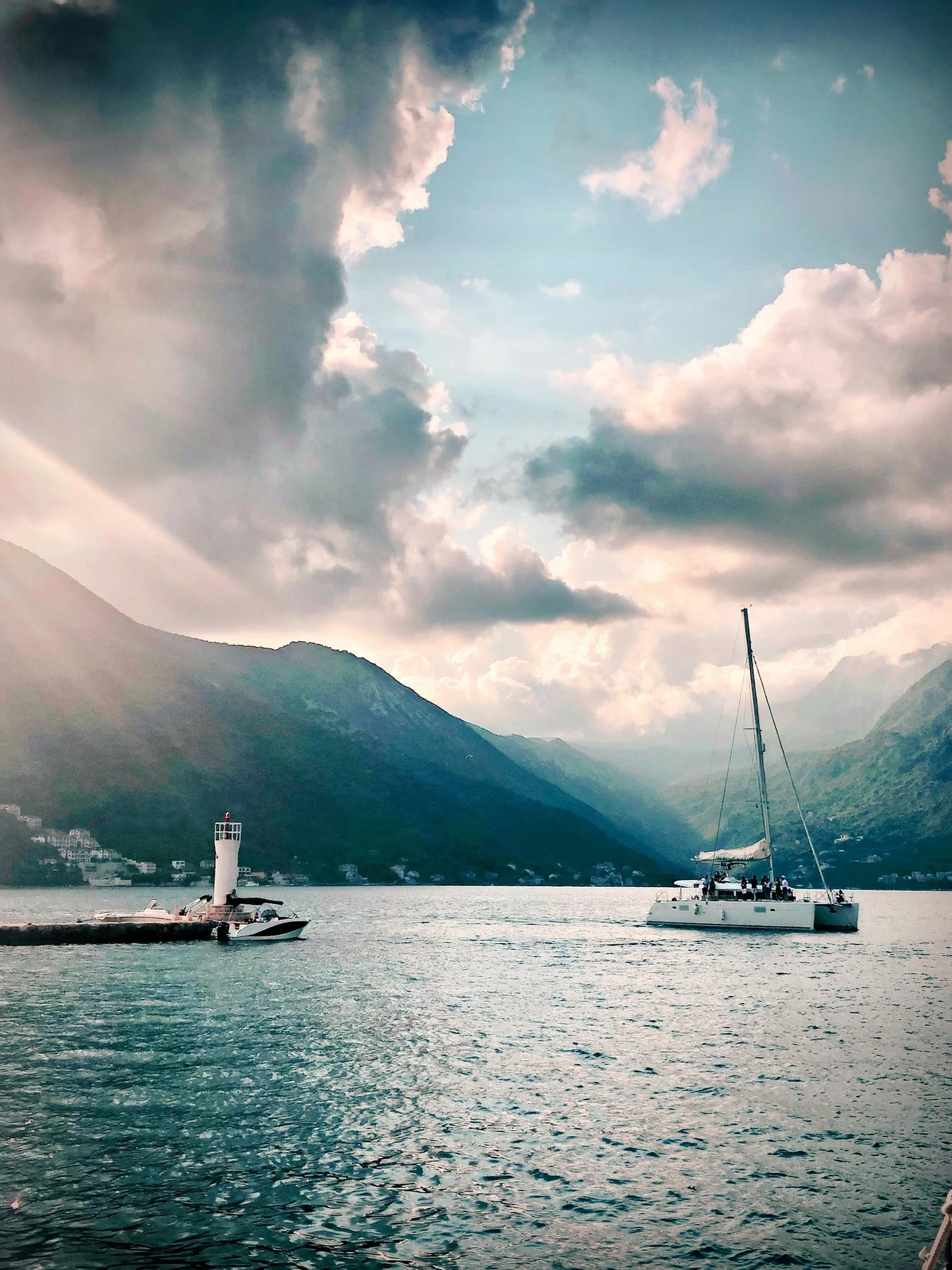 Scenic view of a sailboat on calm water with mountains and cloudy sky in the background, near a small lighthouse on a jetty.