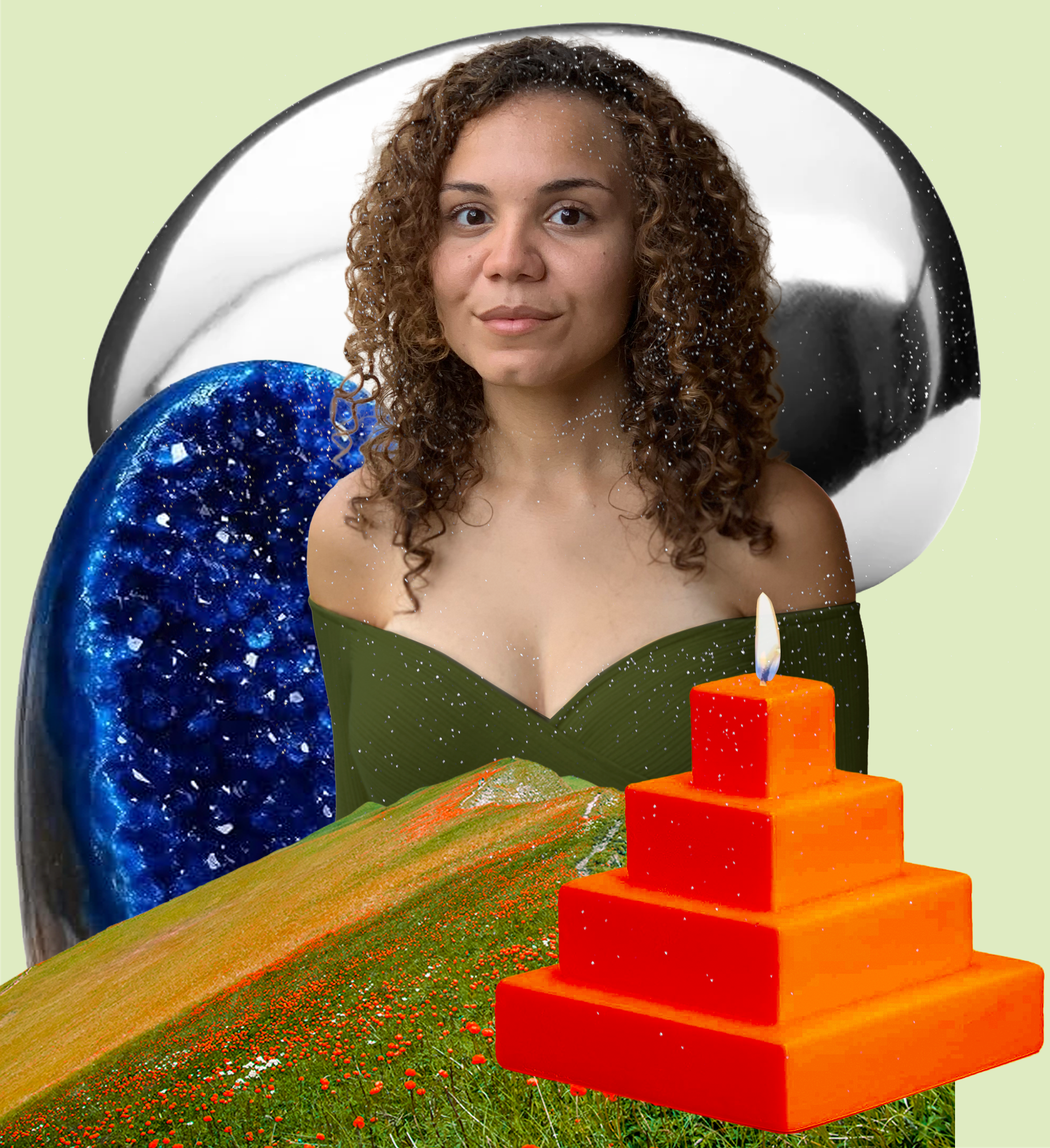 A woman with curly hair sitting on a grassy field with red flowers. In front of her is a small orange stacked cube sculpture with a lit red candle on top of the cubes. There are two large balloons behind her, one blue with shiny speckles and one black and white, creating a colorful scene