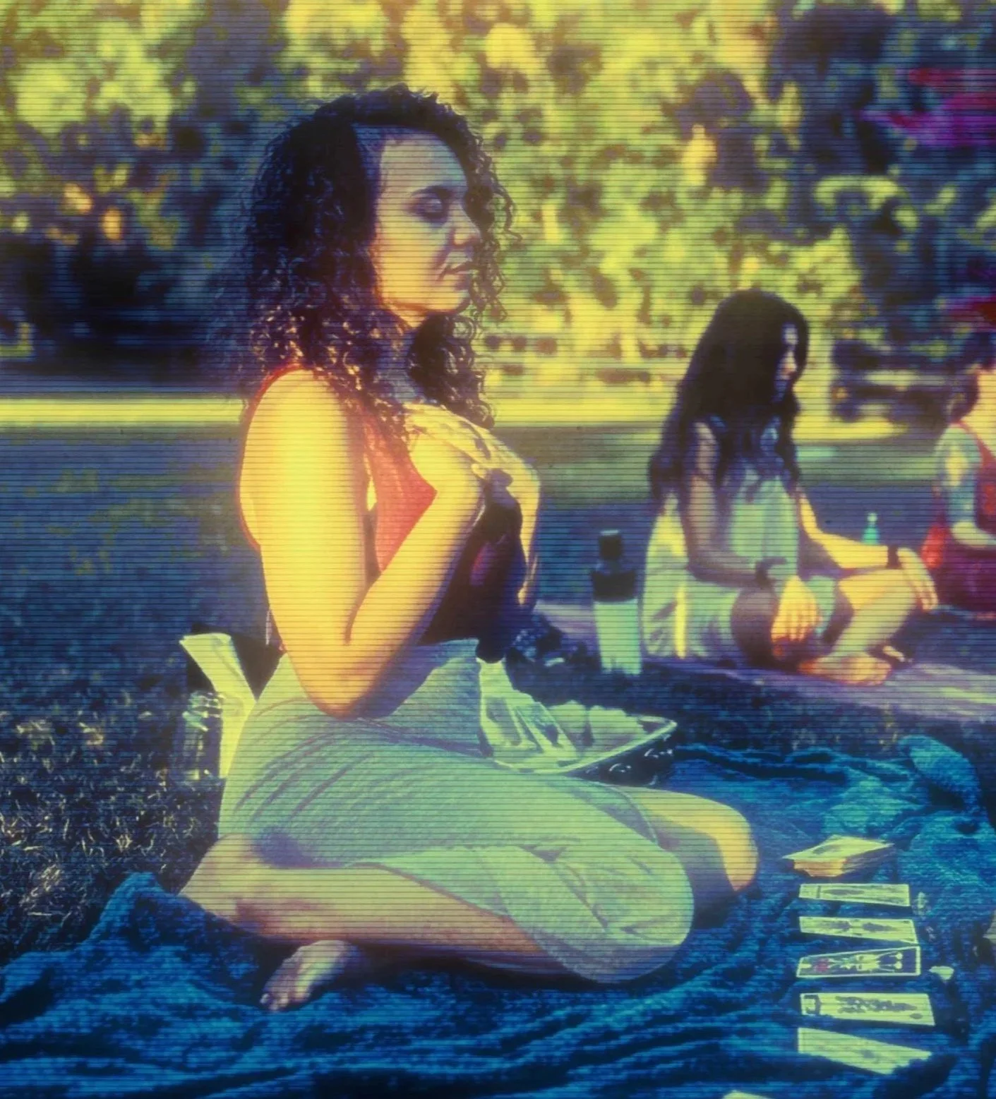A multi-racial woman with curly hair sitting outdoors on a blanket, with a group of people in the background, and tarot cards spread out in front of her as she facilitates a group psychic reading.