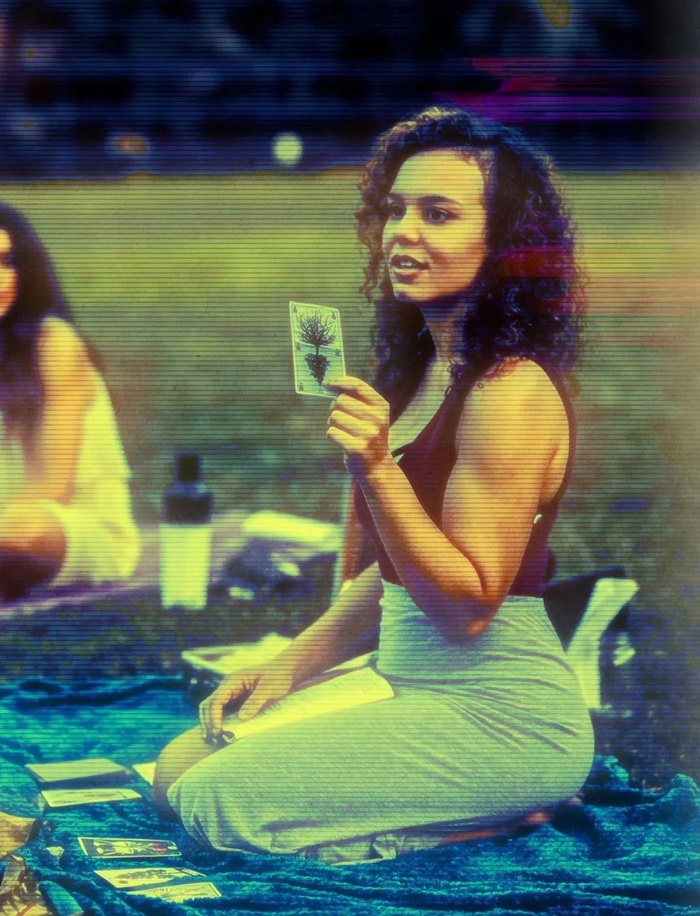 A woman with curly dark hair sitting outdoors at a table, holding a tarot card, with tarot cards and a pink cup in front of her.