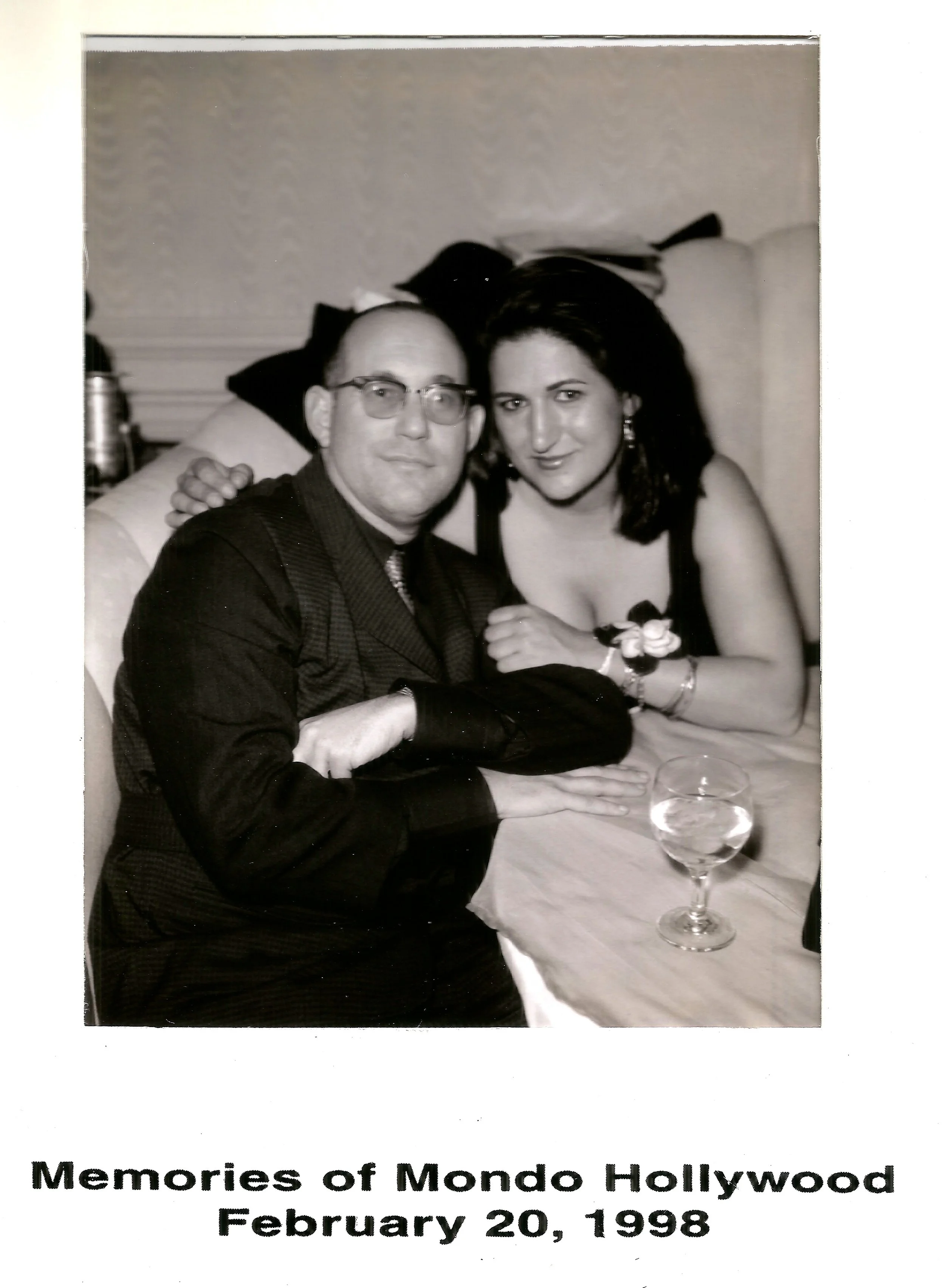 A black-and-white photo of a man and a woman sitting together at a dinner table, with the man wearing glasses and a suit and the woman wearing a dress with earrings, both smiling at the camera. The table has a glass of water in the foreground and some items in the background. The caption at the bottom reads: "Memories of Mondo Hollywood, February 20, 1998."