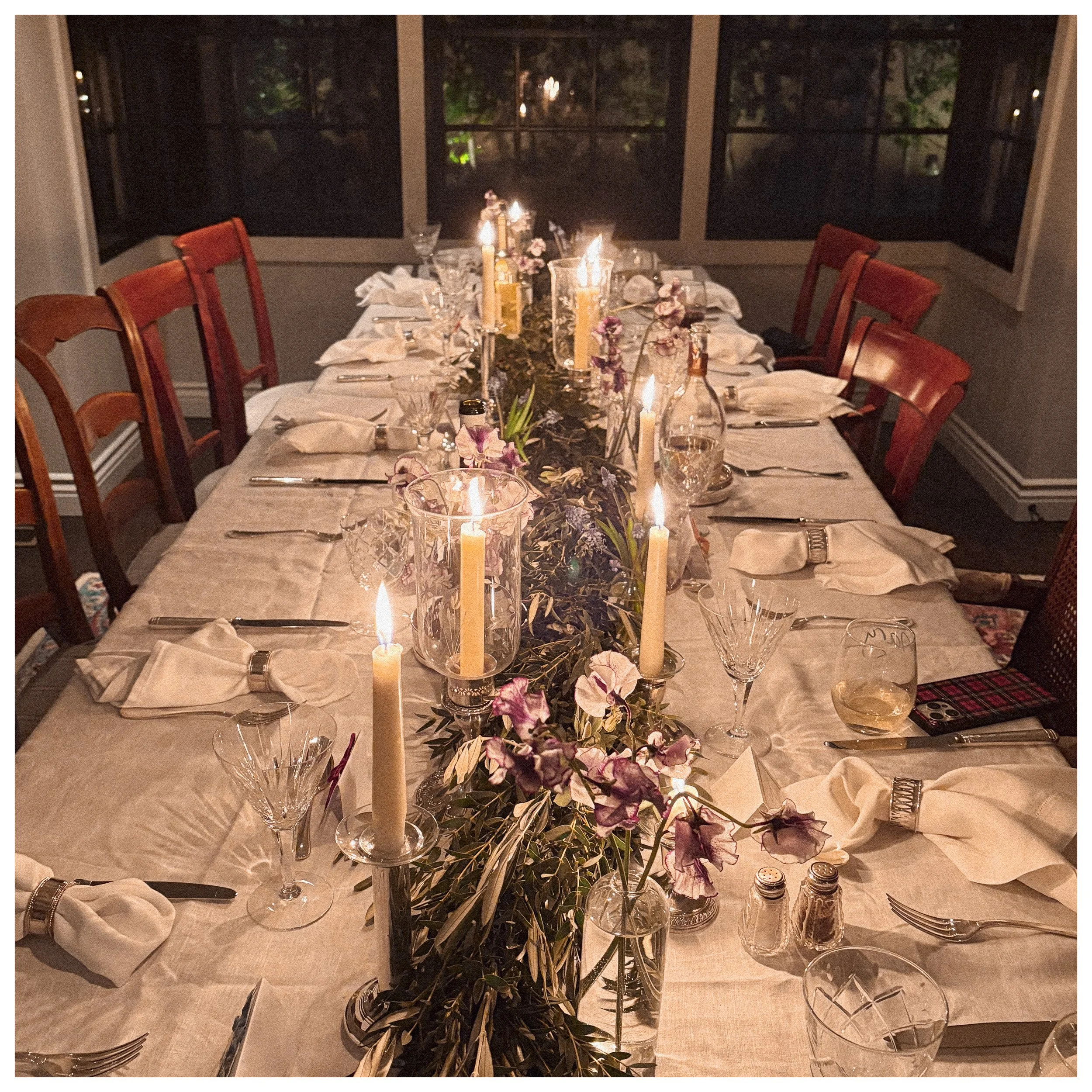 A dining table set for a formal dinner with white napkins, glassware, silverware, and a floral centerpiece with candles. The room has large windows showing a dark outdoor scene.