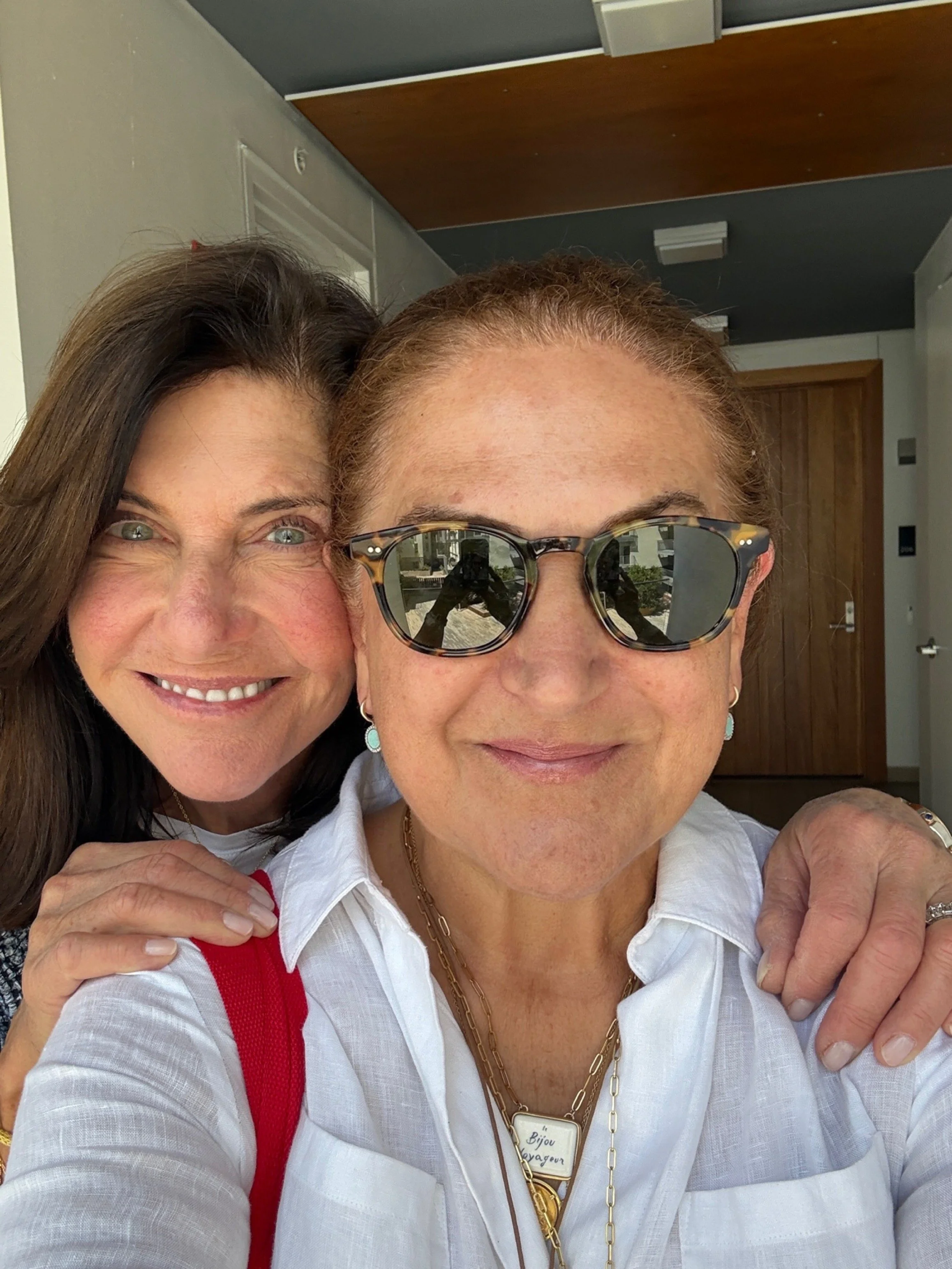 Close-up of two smiling women, one with curly dark hair and the other with short red hair and sunglasses, posed closely together in a bright room.