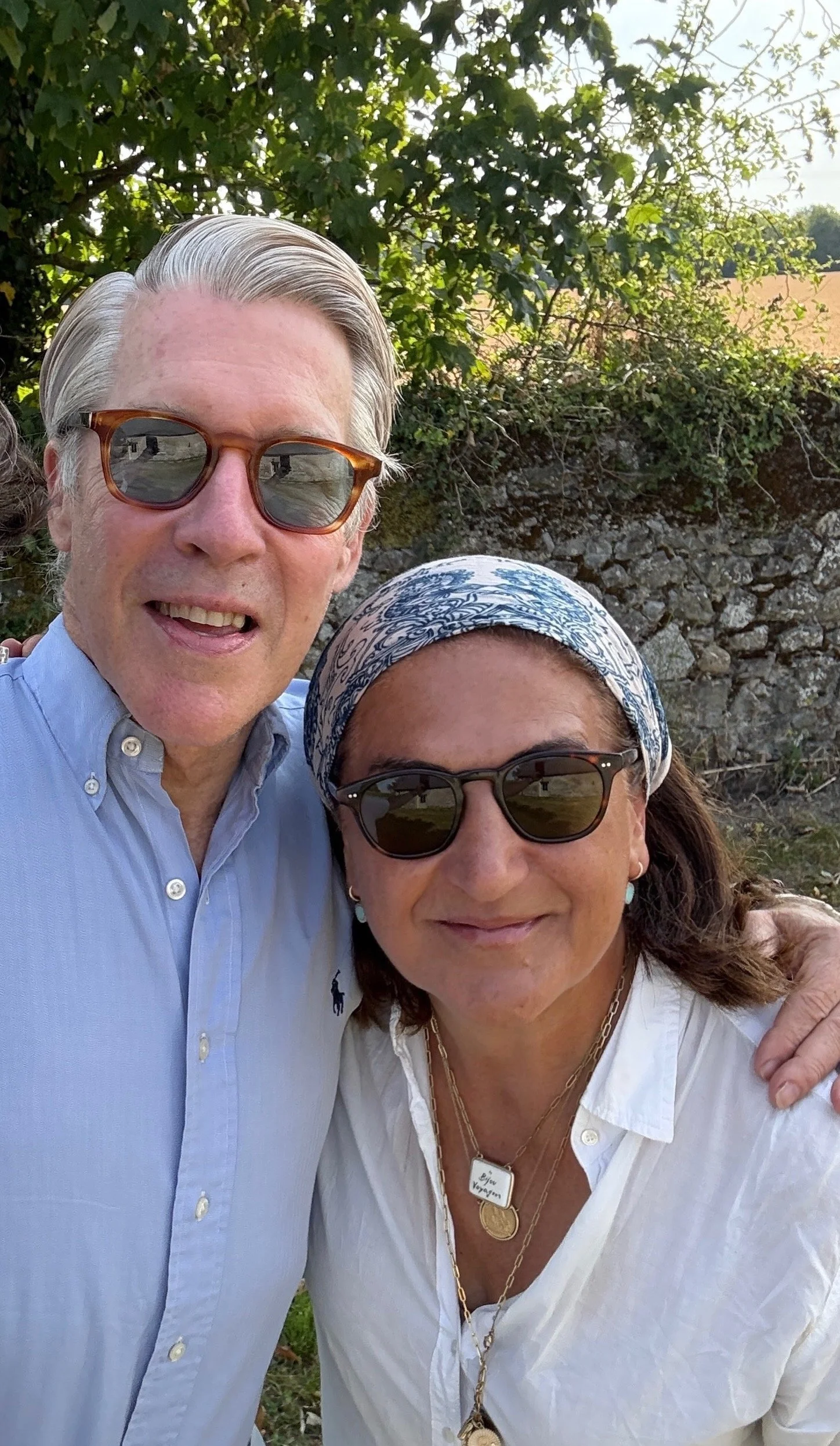 Close-up of a man and woman outdoors, both wearing sunglasses and smiling, with greenery and rocks in the background.