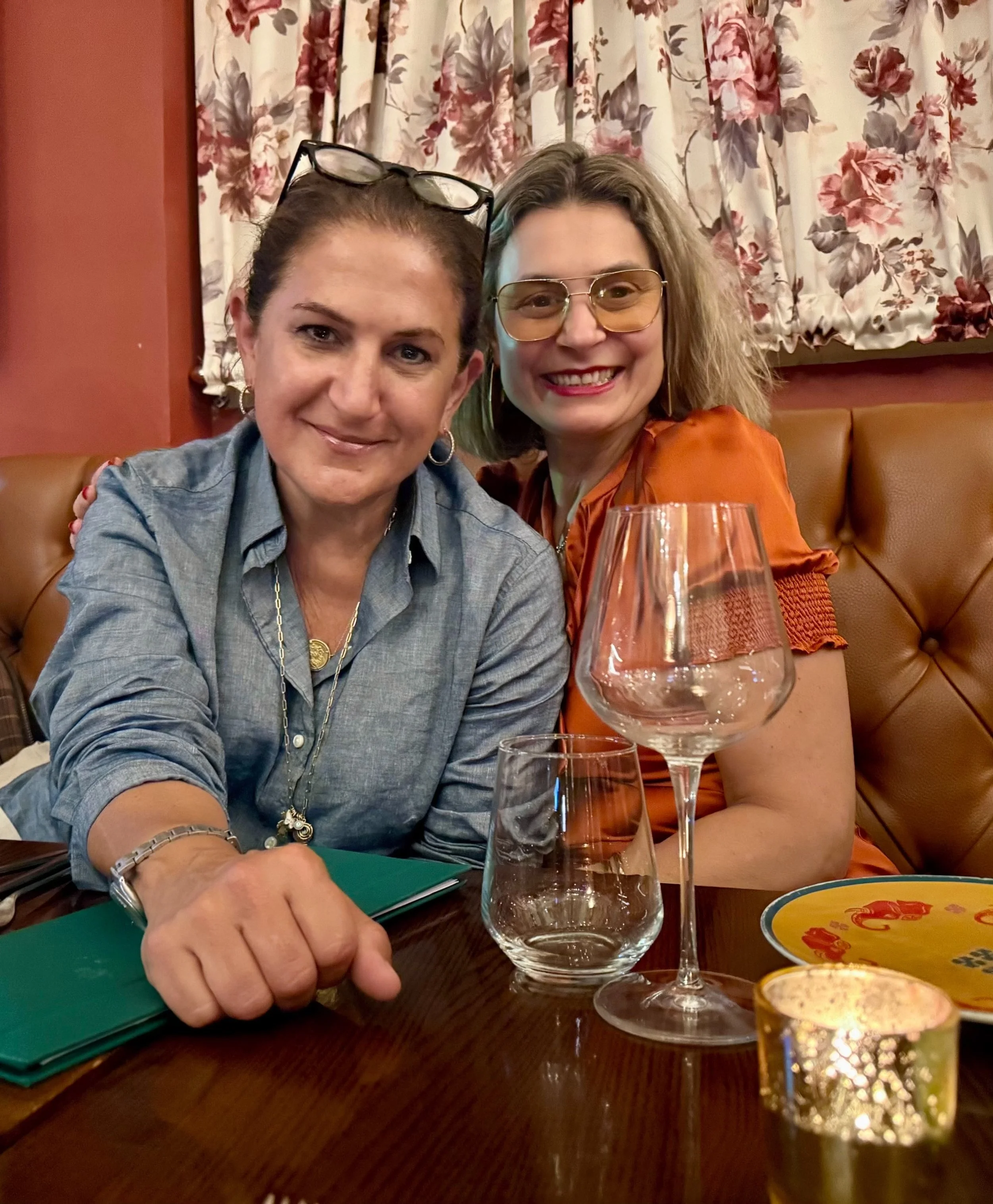 Two women sitting at a restaurant table, smiling for a selfie. The woman on the left has dark hair, glasses on her head, and is wearing a denim shirt. The woman on the right has curly blonde hair, glasses, and an orange outfit. There are empty wine glasses on the table and a decorative candle holder in the foreground. The background shows floral curtains and a brown wall.