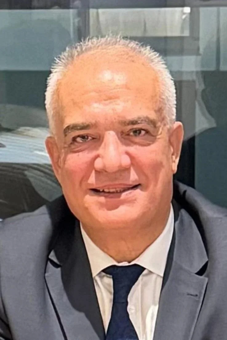 Close-up of a smiling elderly man in a gray suit and tie, with a glass window and office environment in the background.