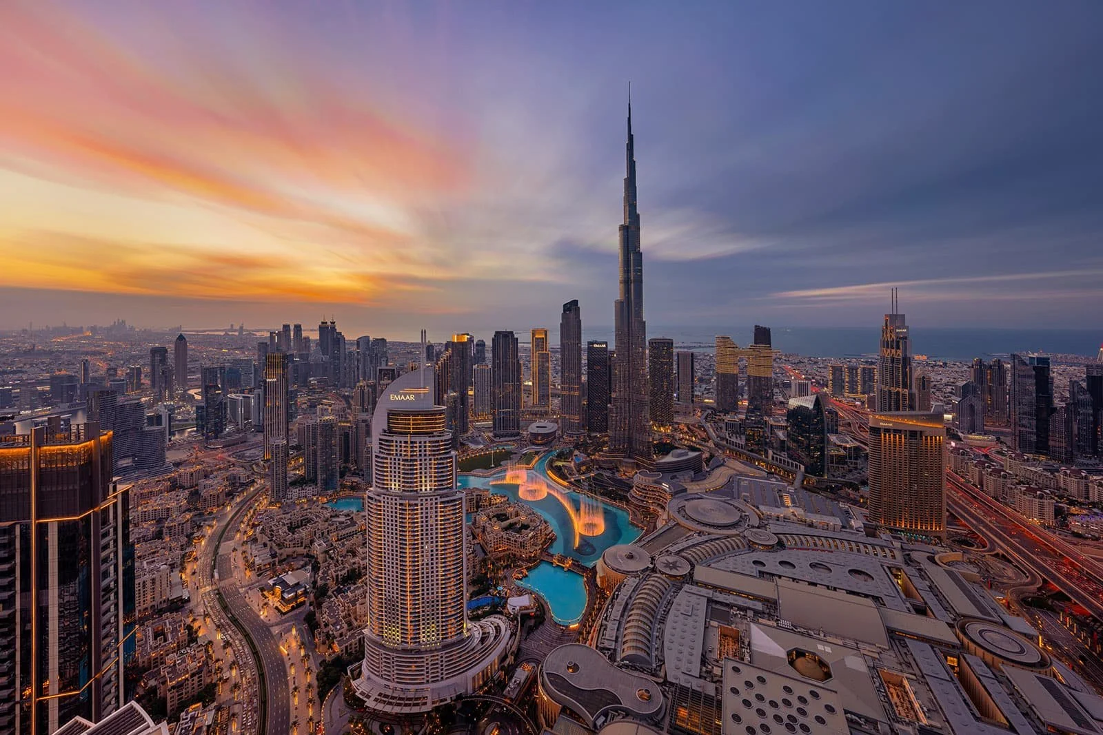 A city skyline at sunset with tall skyscrapers, including the Burj Khalifa, and a large artificial lake with fountains.