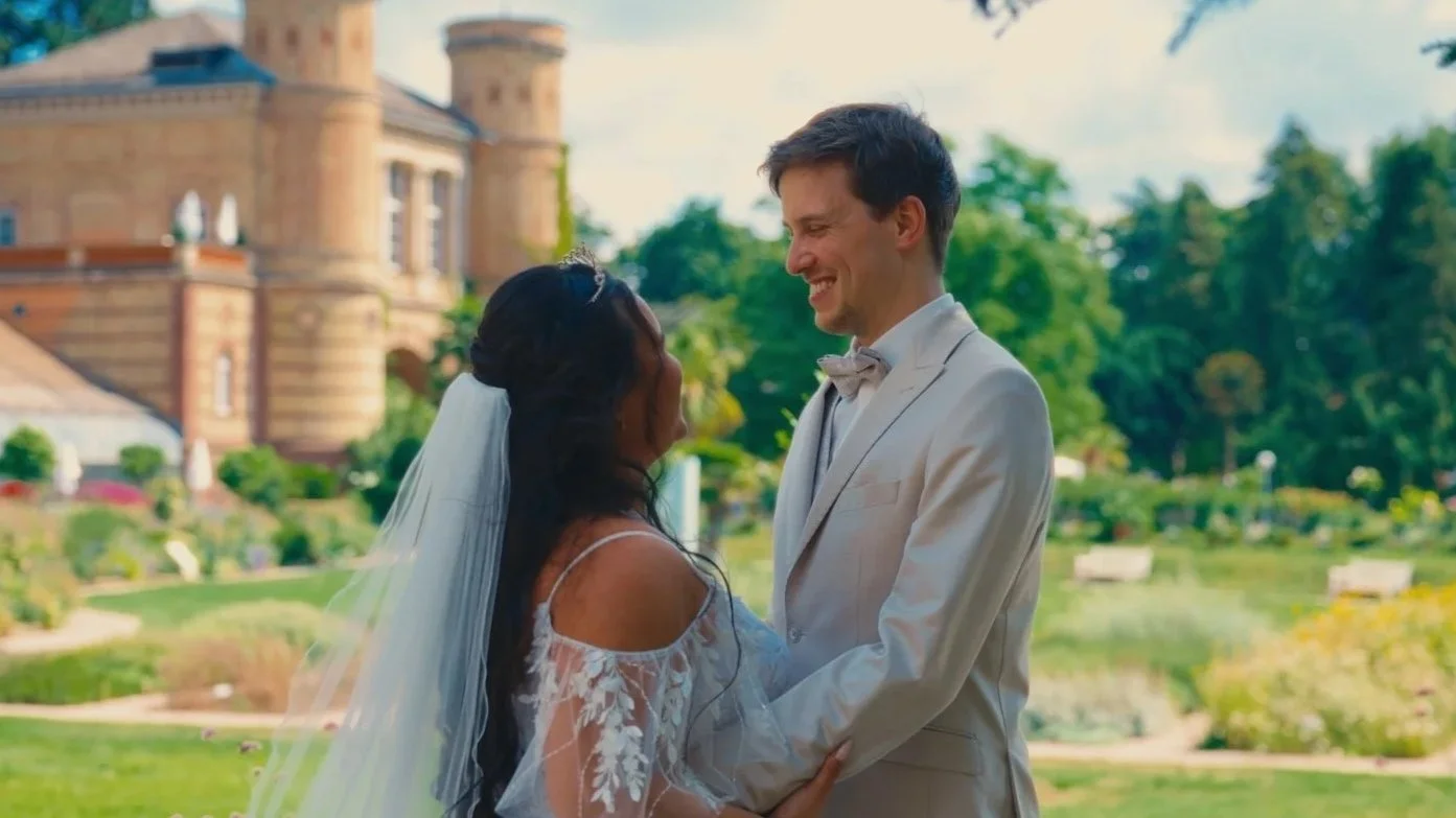 A couple in wedding attire standing outdoors in front of a castle, smiling at each other during a wedding ceremony.