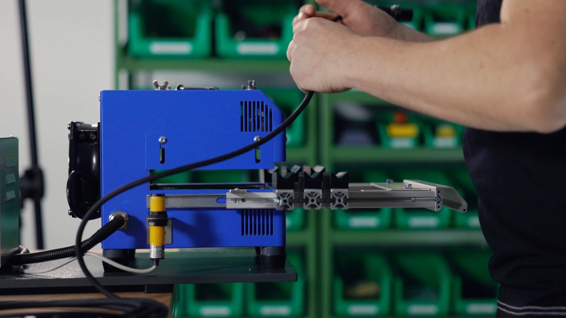 Person working with electronic equipment inside a workshop with green storage bins in the background.