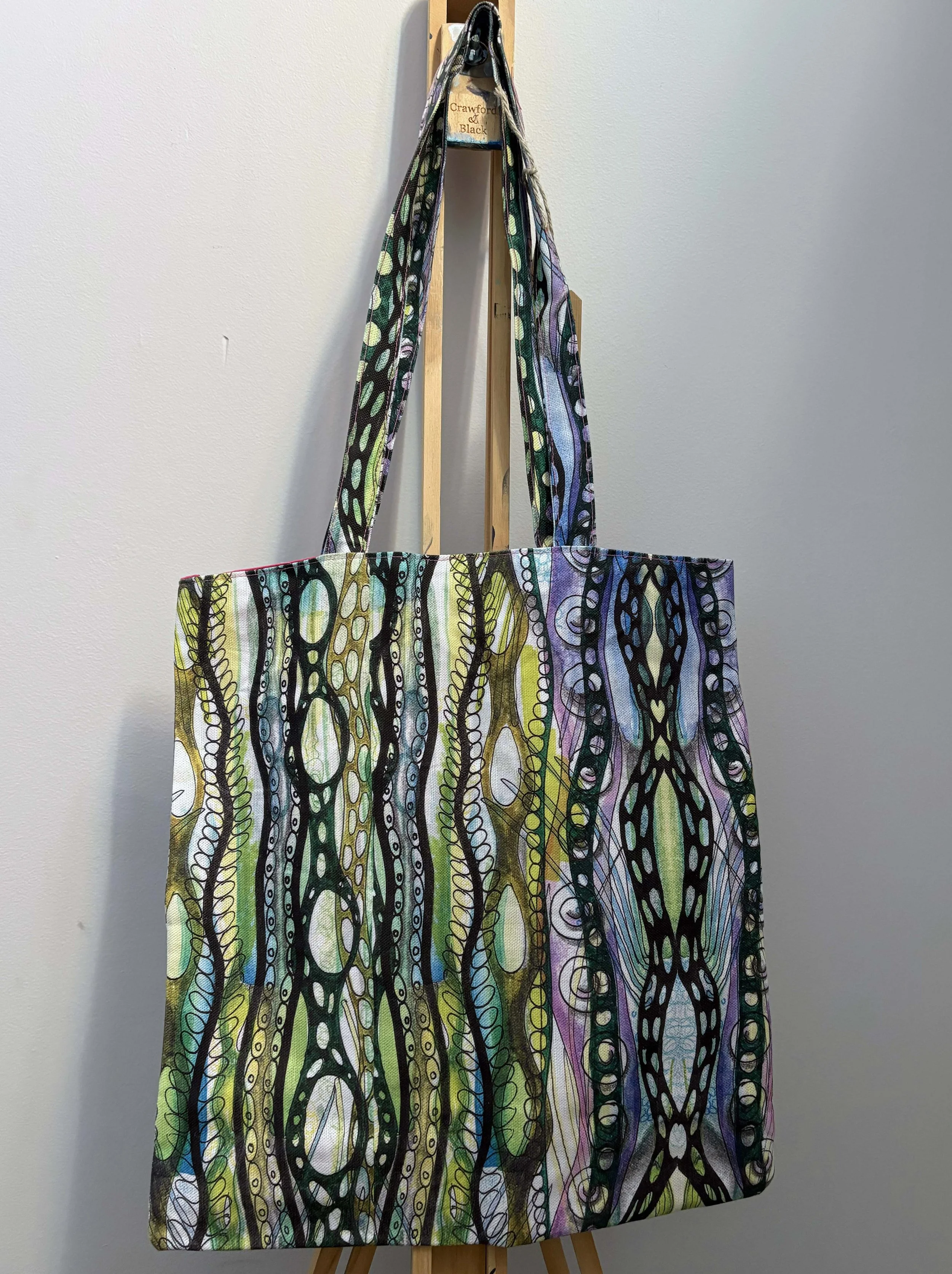 A colorful tote bag with an abstract pattern of black, green, purple, and blue lines and shapes, hanging on a wooden easel against a plain wall.