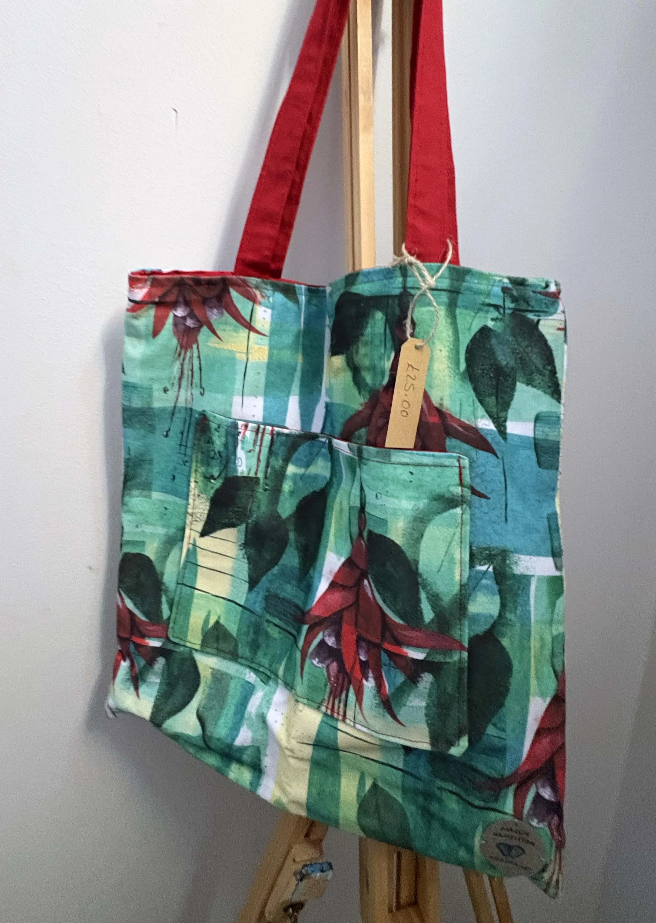 A colorful fabric tote bag with a botanical print of green leaves and red flowers, hanging on a wooden easel. The bag has red handles, a front pocket, and a price tag showing $25.00.