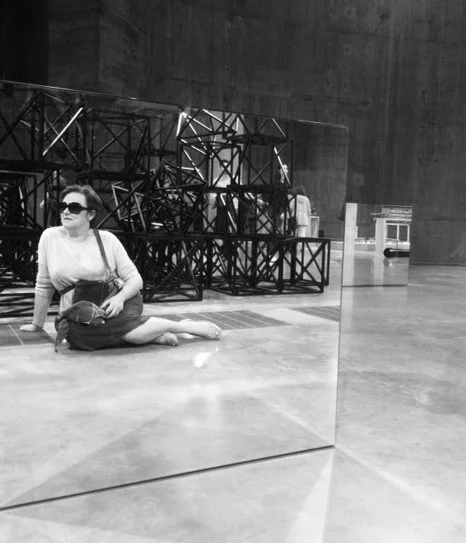Woman sitting on the floor in a modern art gallery, with a reflection in a mirrored surface, surrounded by abstract metal structures and sculptures.