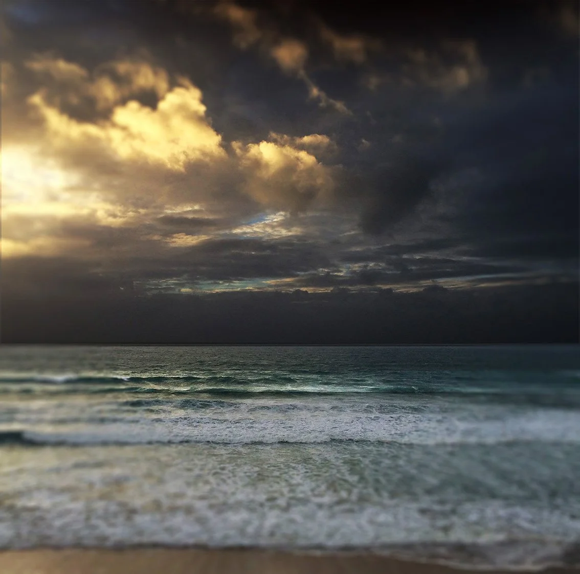 Dark, cloudy sky over a calm ocean at sunset with waves gently reaching the shore.