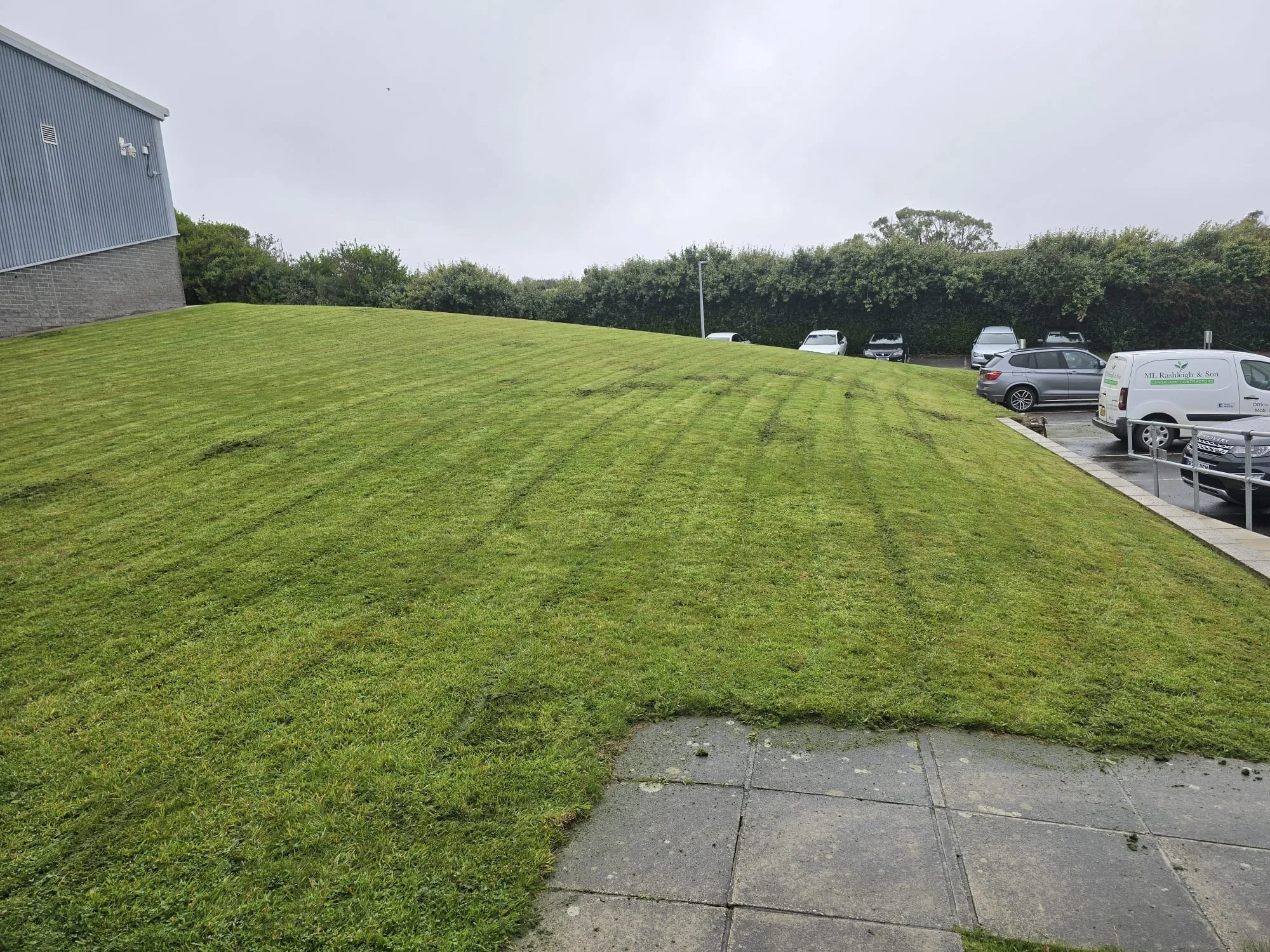 Lawn mower on freshly mowed green grass overlooking the Helford river with boats, trees, and a patio with outdoor furniture in the background.