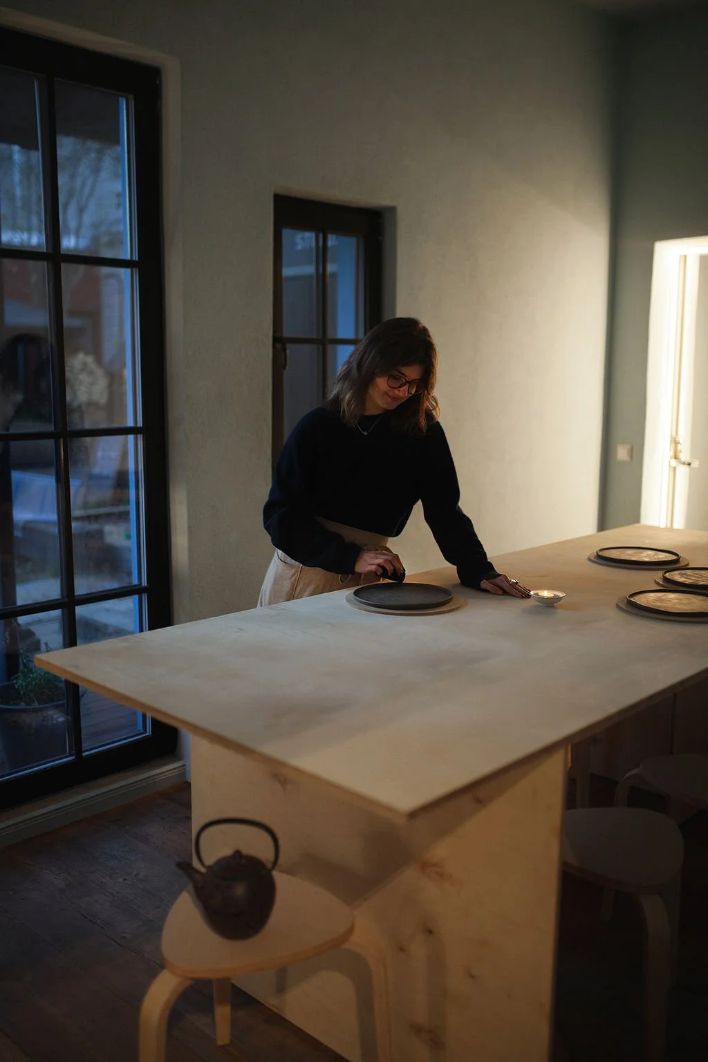 A woman with glasses and shoulder-length hair, wearing a black sweater and beige pants, lighting a candle on a wooden kitchen island with black plates. The room has large windows and a door, with a teapot on a small round stool in front of the island.