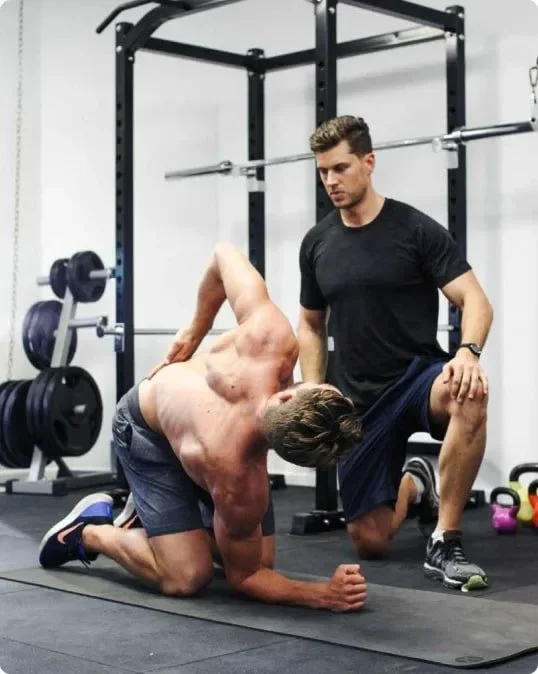 A personal trainer helps a muscular man stretch during a workout in a gym with weightlifting equipment.