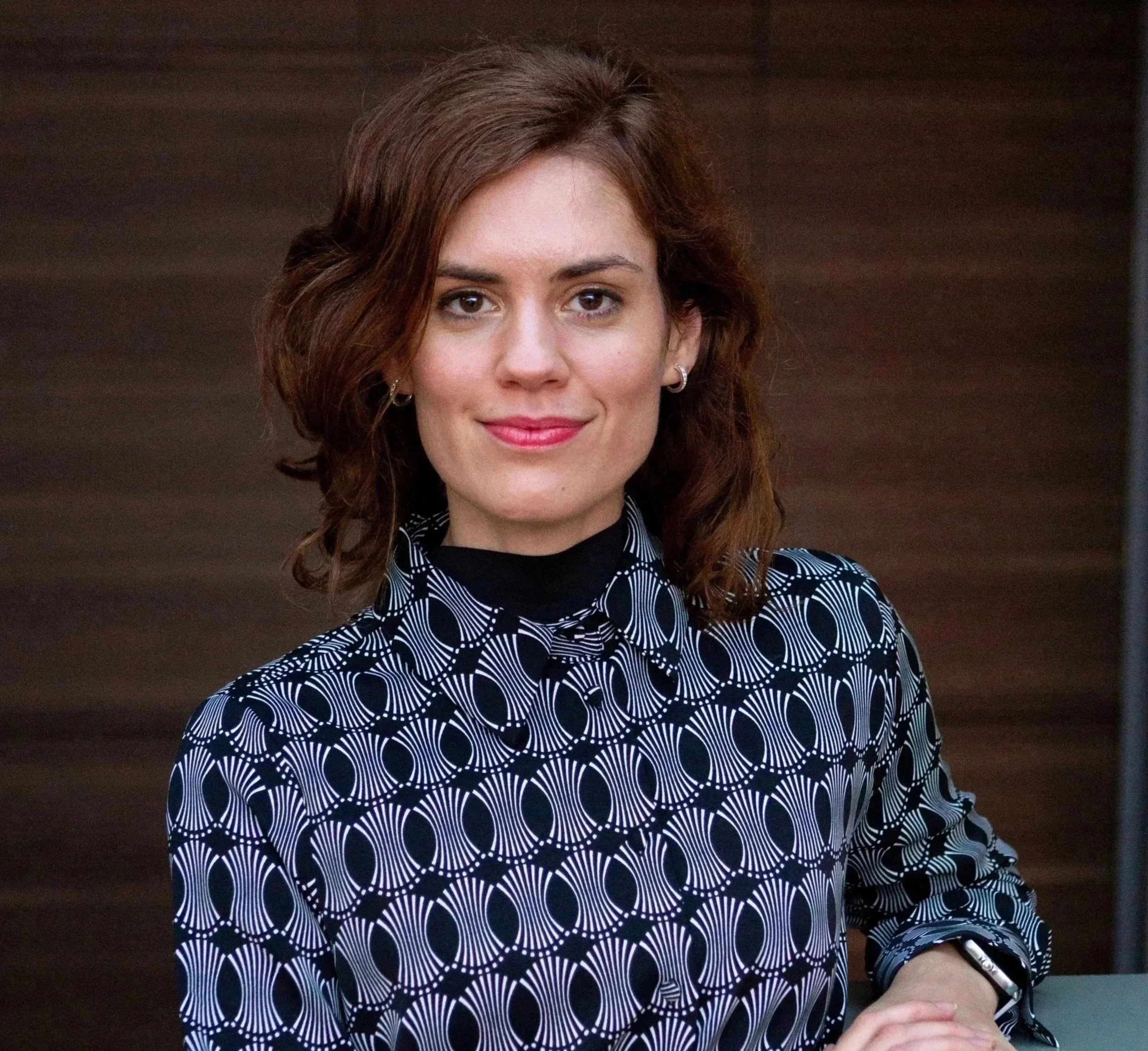A woman with dark, wavy shoulder-length hair smiling, wearing hoop earrings, a black top, and a mint green blazer, in an indoor setting.