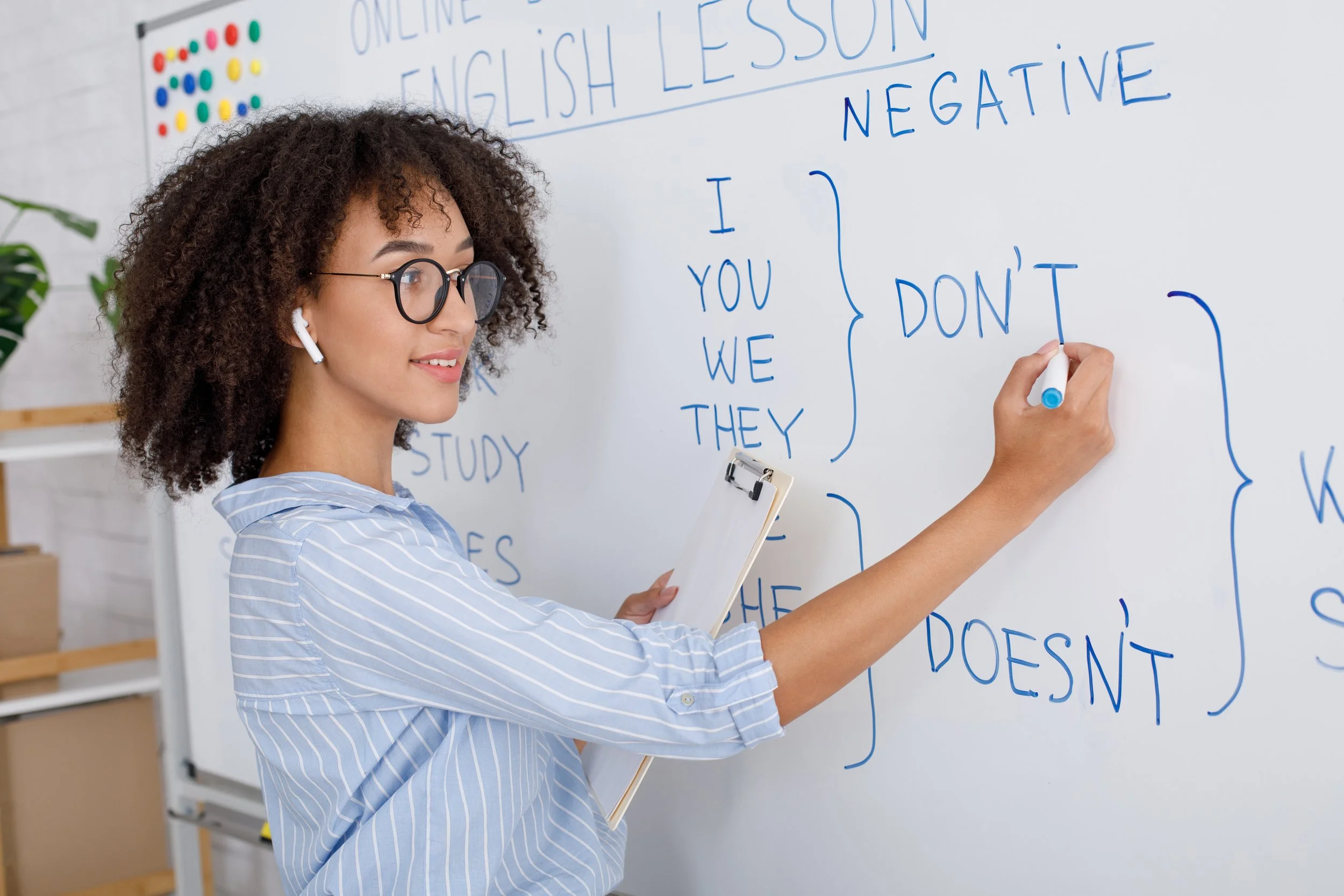 A woman teaching an online English lesson stands at a whiteboard explaining negative contractions, such as "don't" and "doesn't." She is holding a clipboard and writing with a blue marker.