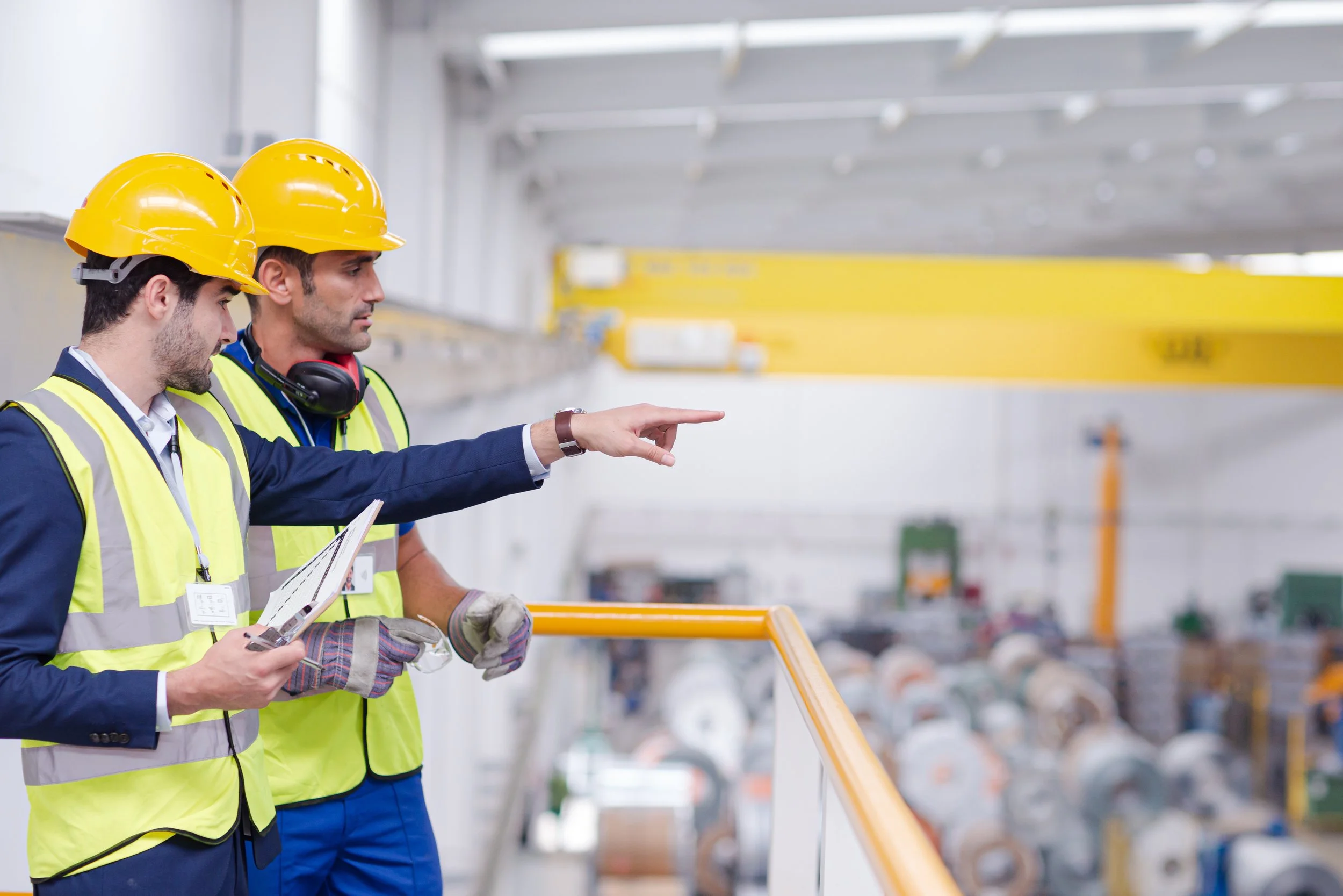 Two workers in hard hats and reflective vests discussing in a factory setting.
