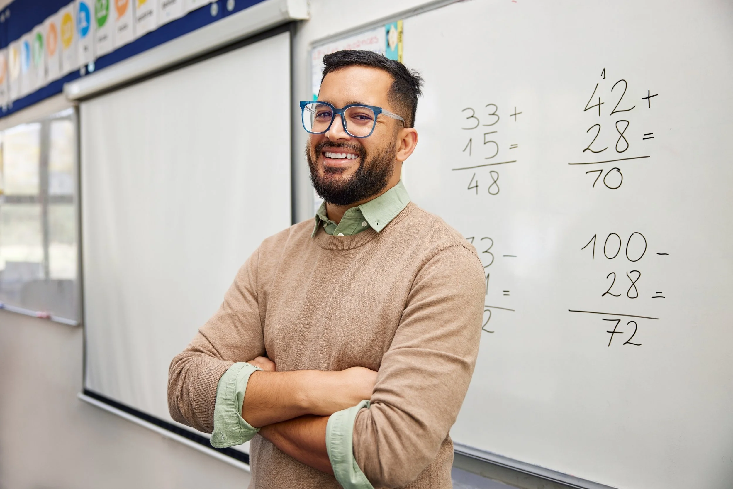 A teacher with glasses and a beard stands smiling in front of a whiteboard with math problems written on it.