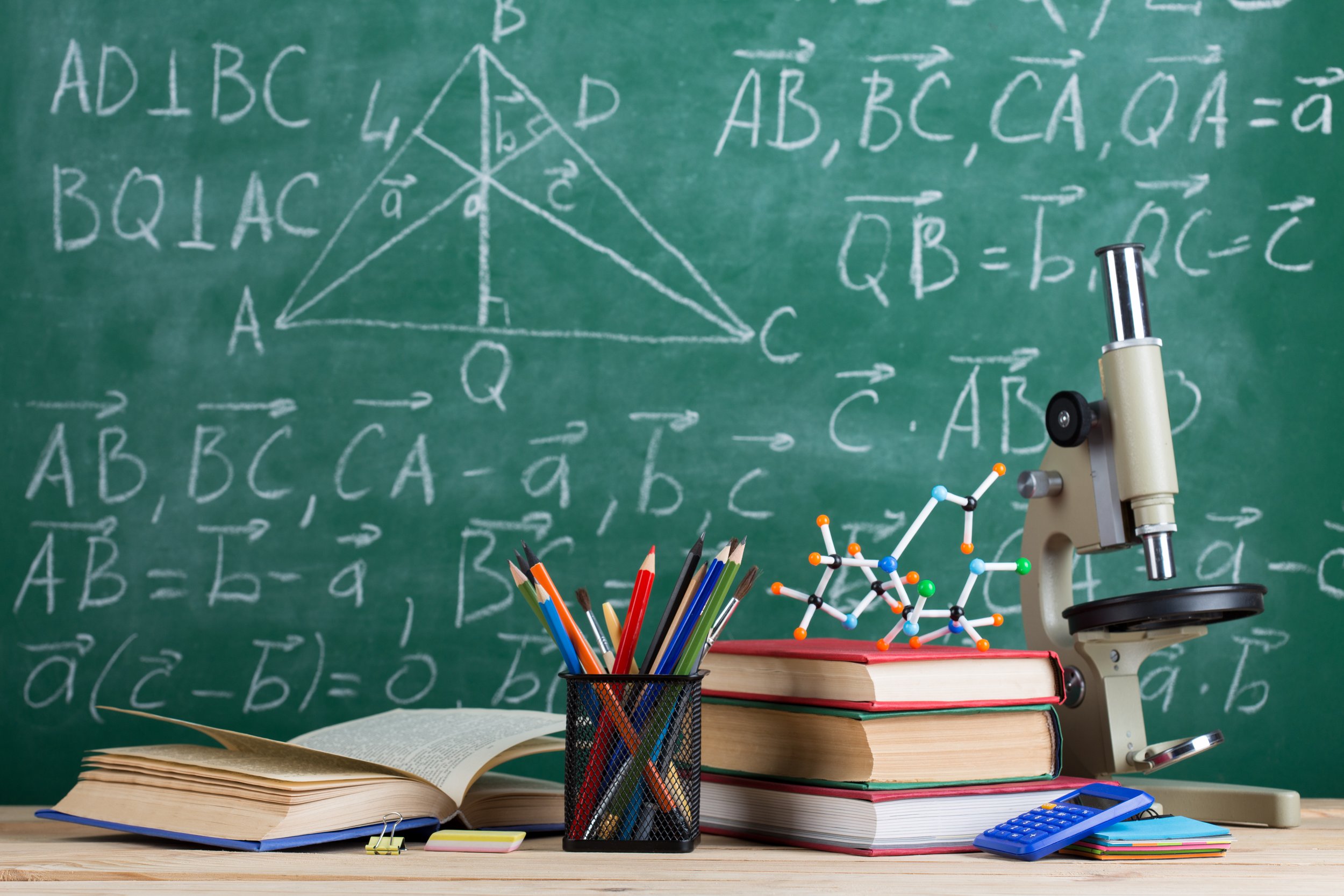School supplies and a microscope on a desk in front of a chalkboard with math equations.