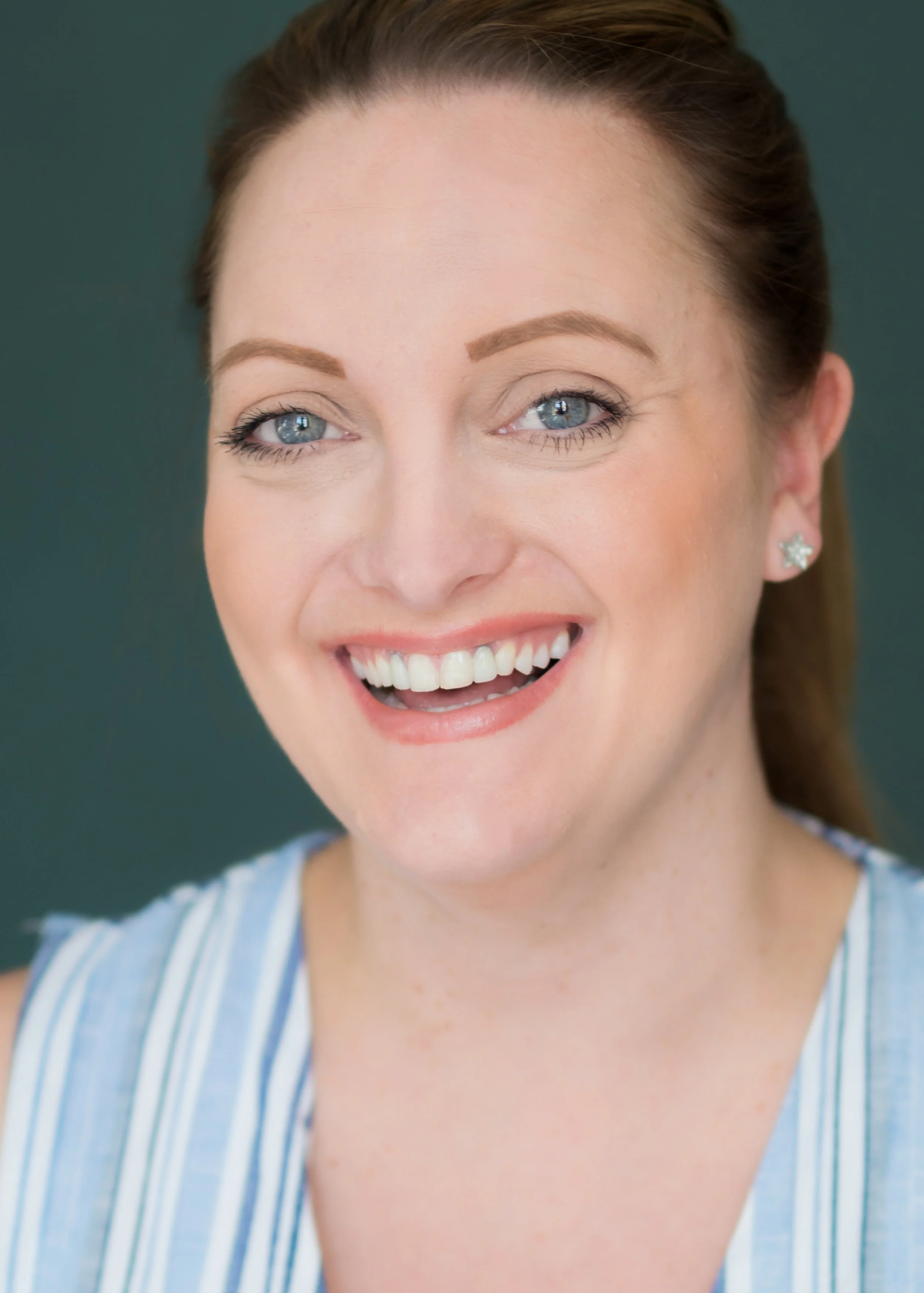 A smiling woman with blue eyes, light skin, red hair pulled back, wearing star-shaped earrings and a blue and white striped top.