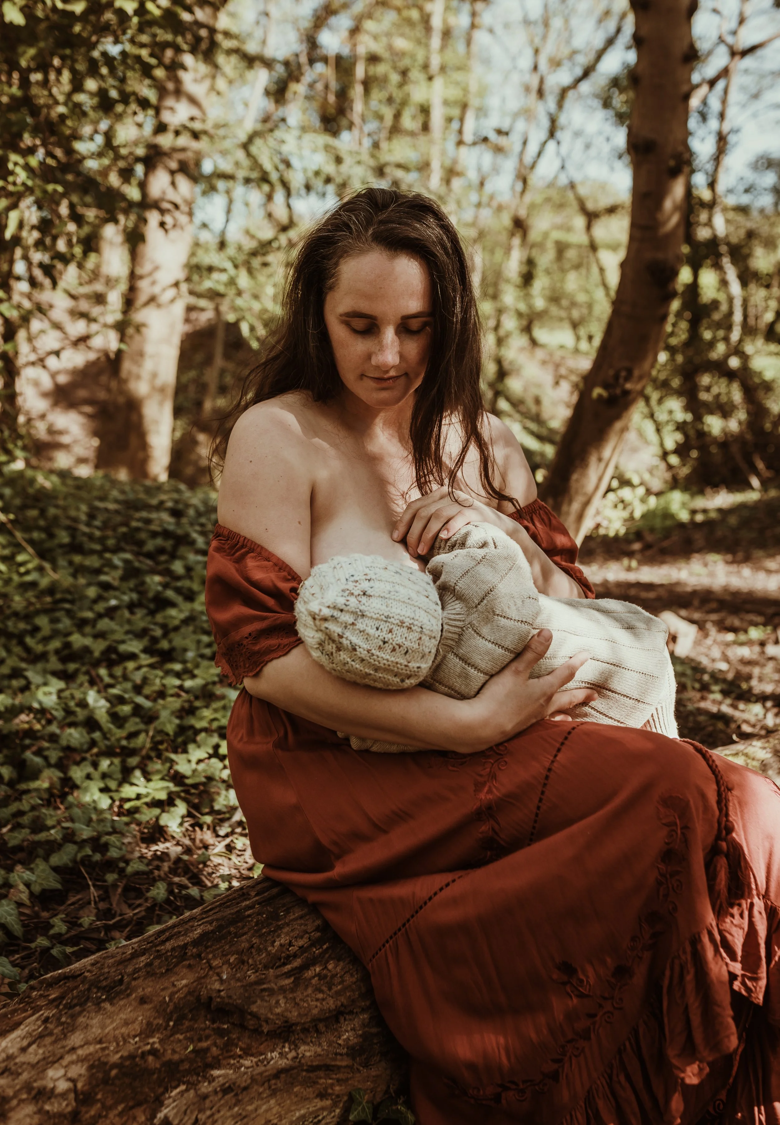Mother nursing her baby in a rust red dress in Scottish woodland during an outdoor nursing photography session with Marieke Captures Birth