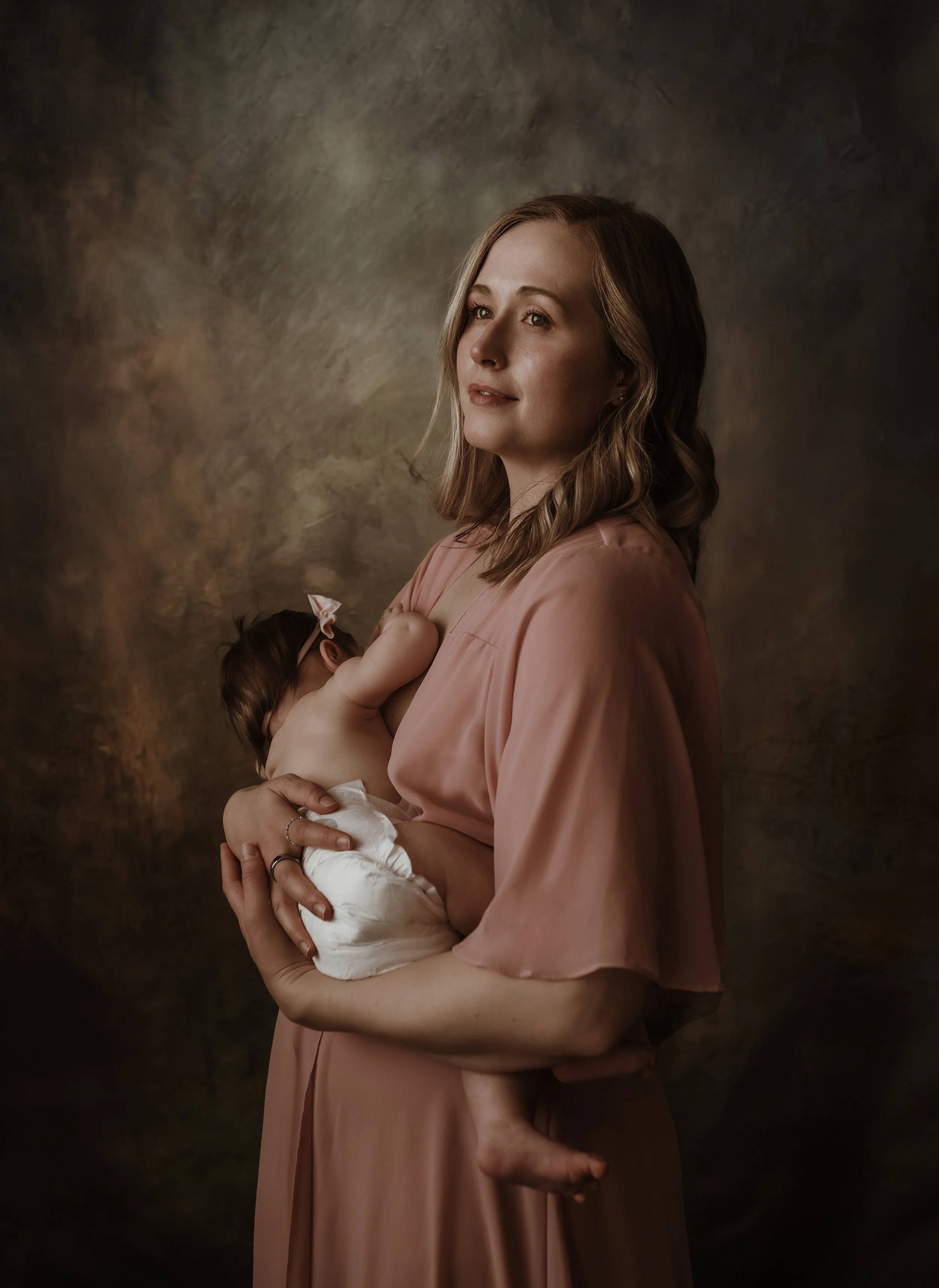 Studio portrait of a mother holding her baby in a soft pink dress against a moody backdrop during a nursing photography session with Marieke Captures Birth Glasgow