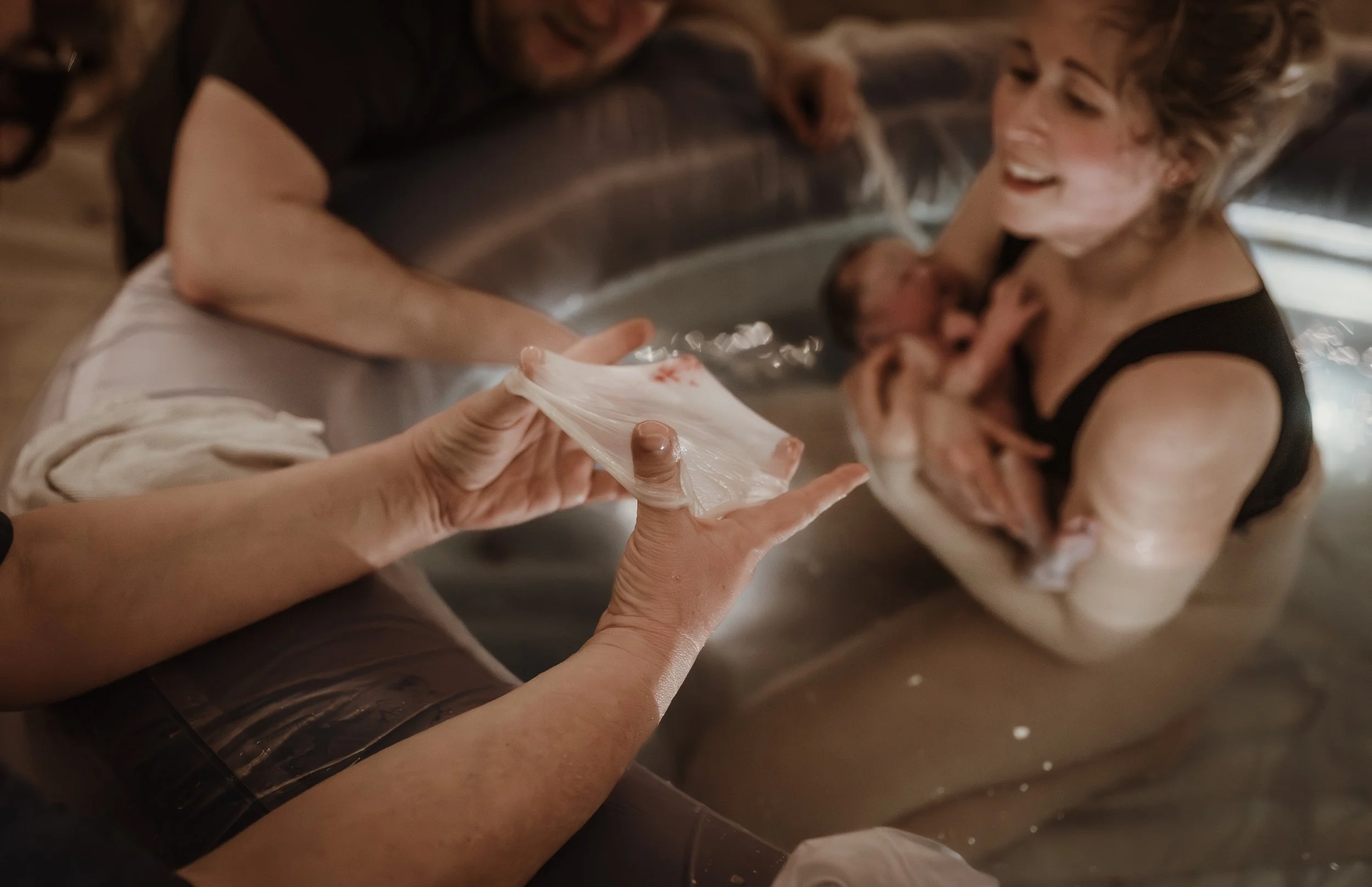 Midwife holding up the intact amniotic sac after a Glasgow home water birth, with Rebekah and James visible in the birth pool in the background