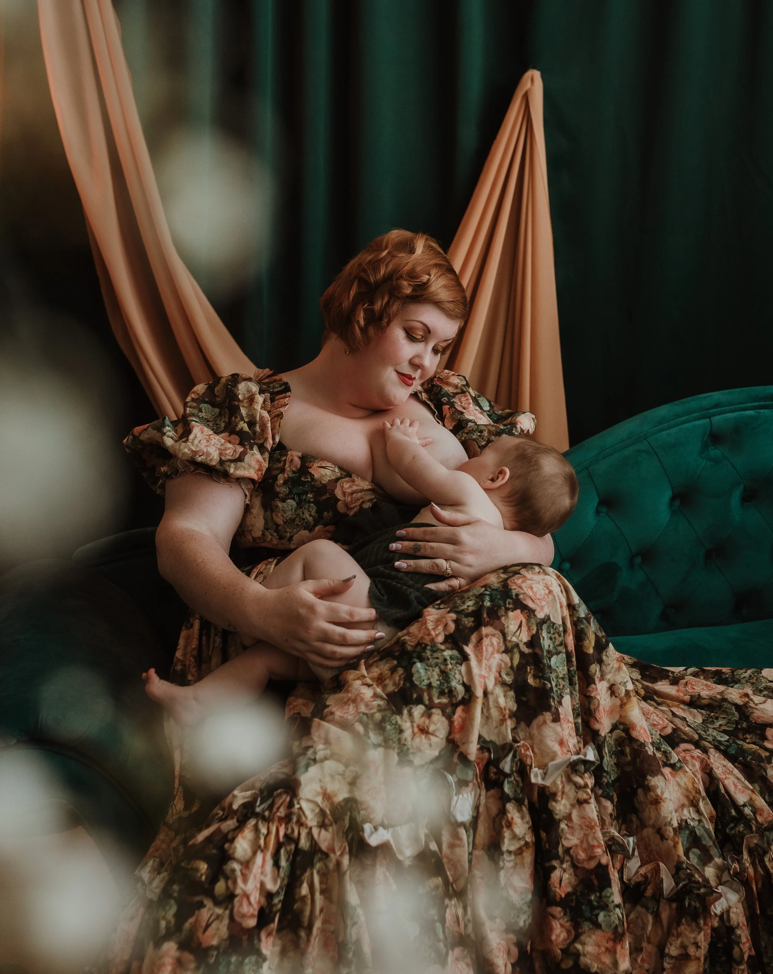 Mother nursing her baby in a floral dress on a dark velvet sofa during a studio nursing photography session with Glasgow photographer Marieke Captures Birth
