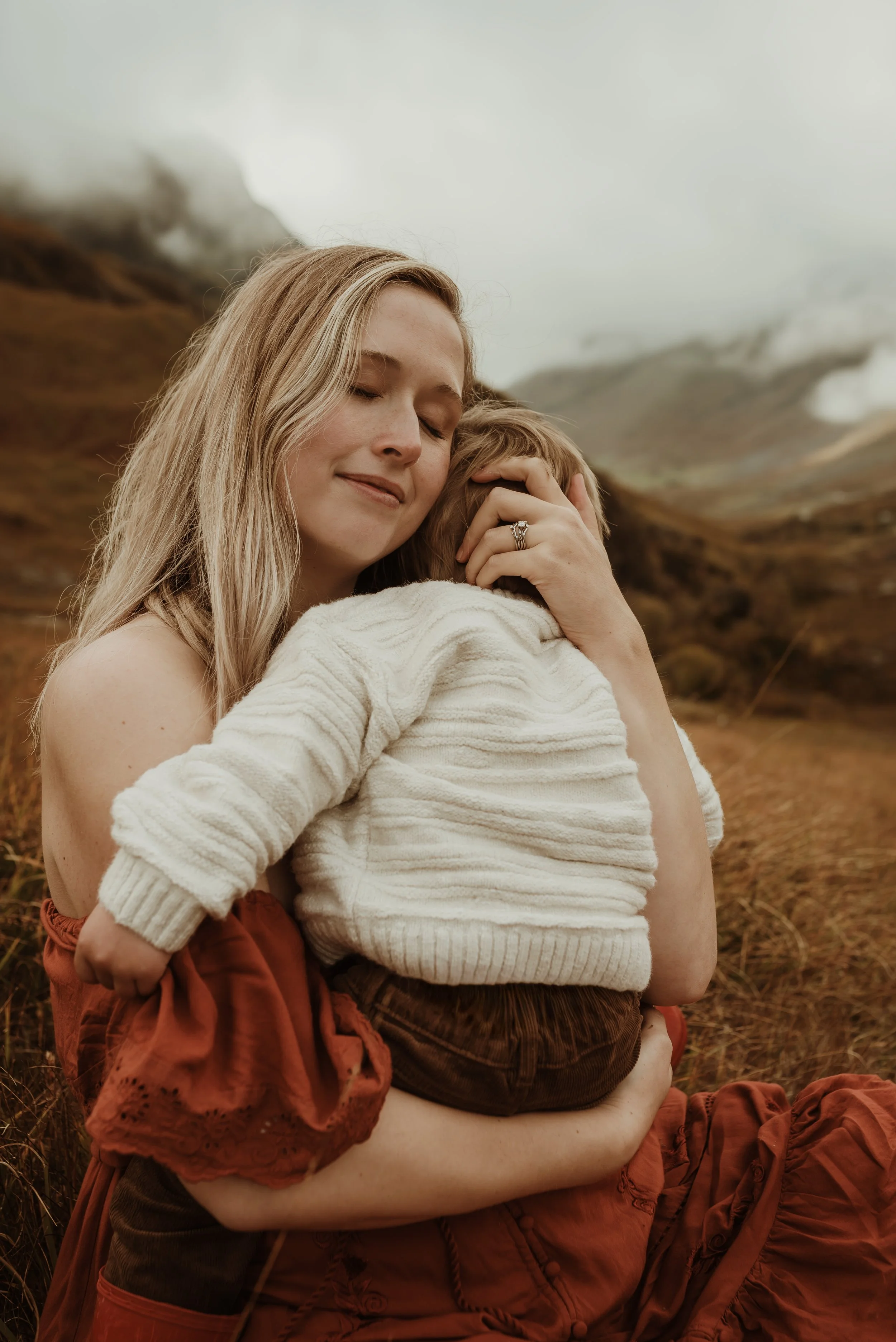 Expectant mother walking through the misty mountains of Glencoe during an adventurous maternity session in Scotland, captured by a Glasgow birth photographer as her toddler runs ahead in red wellies.