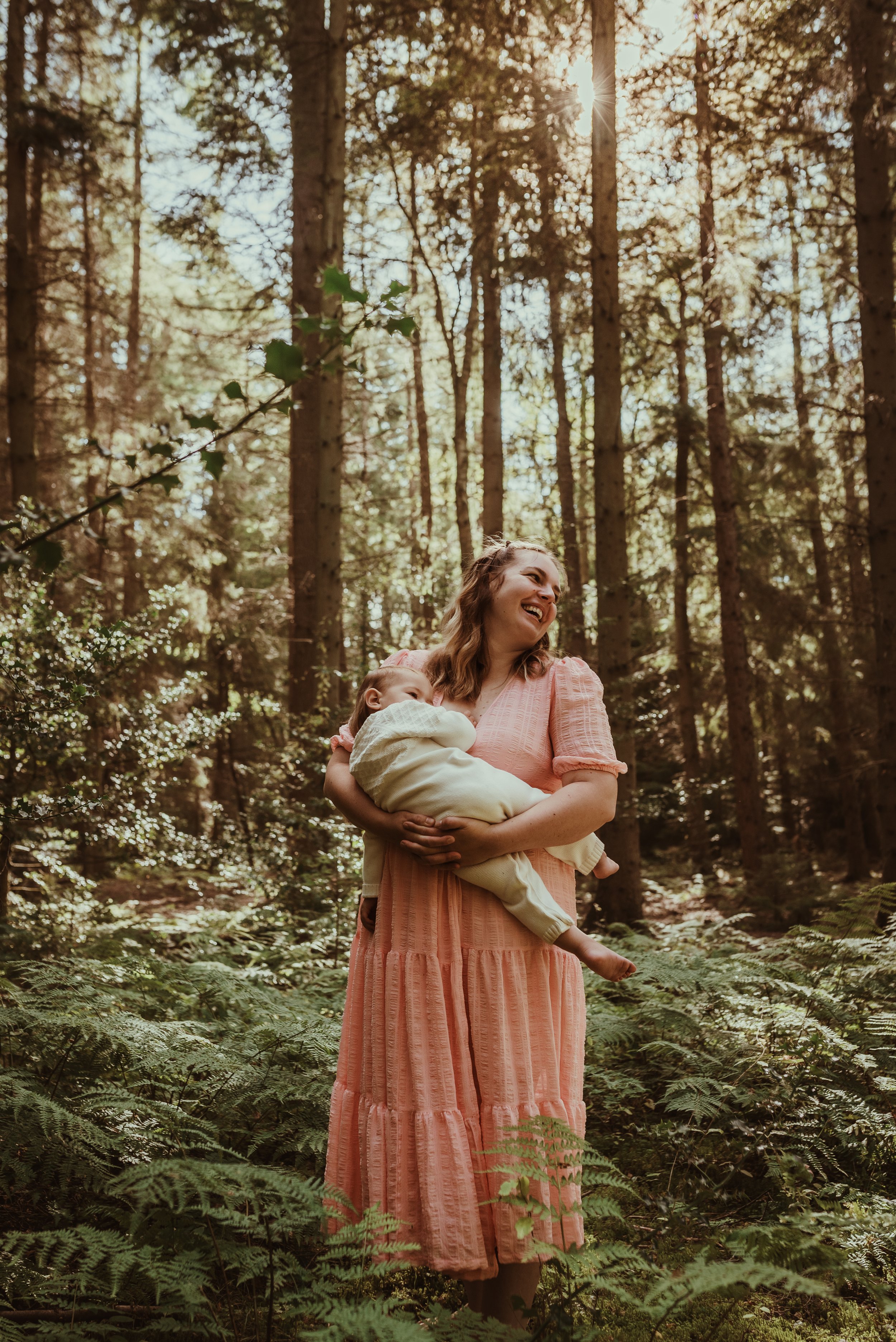 Mother standing in Scottish woodland holding her baby in a pink dress during an outdoor nursing photography session with Marieke Captures Birth