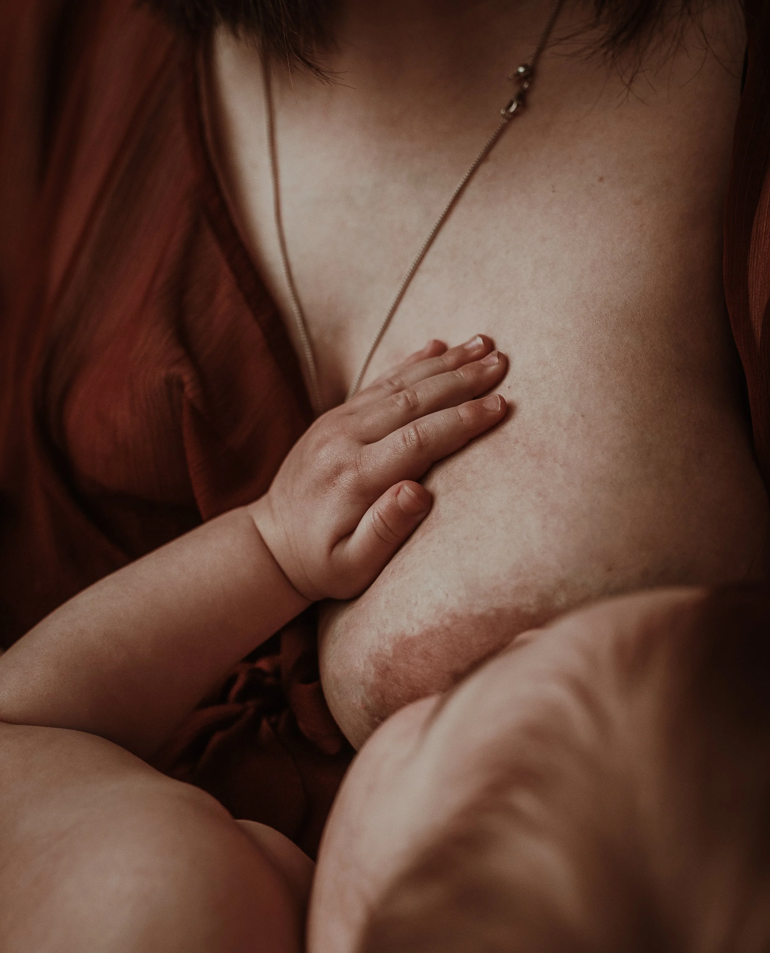 A close-up detail photograph from a nursing photography session in Glasgow, showing a baby's small hand resting gently on their mother's chest while breastfeeding, warm tones throughout