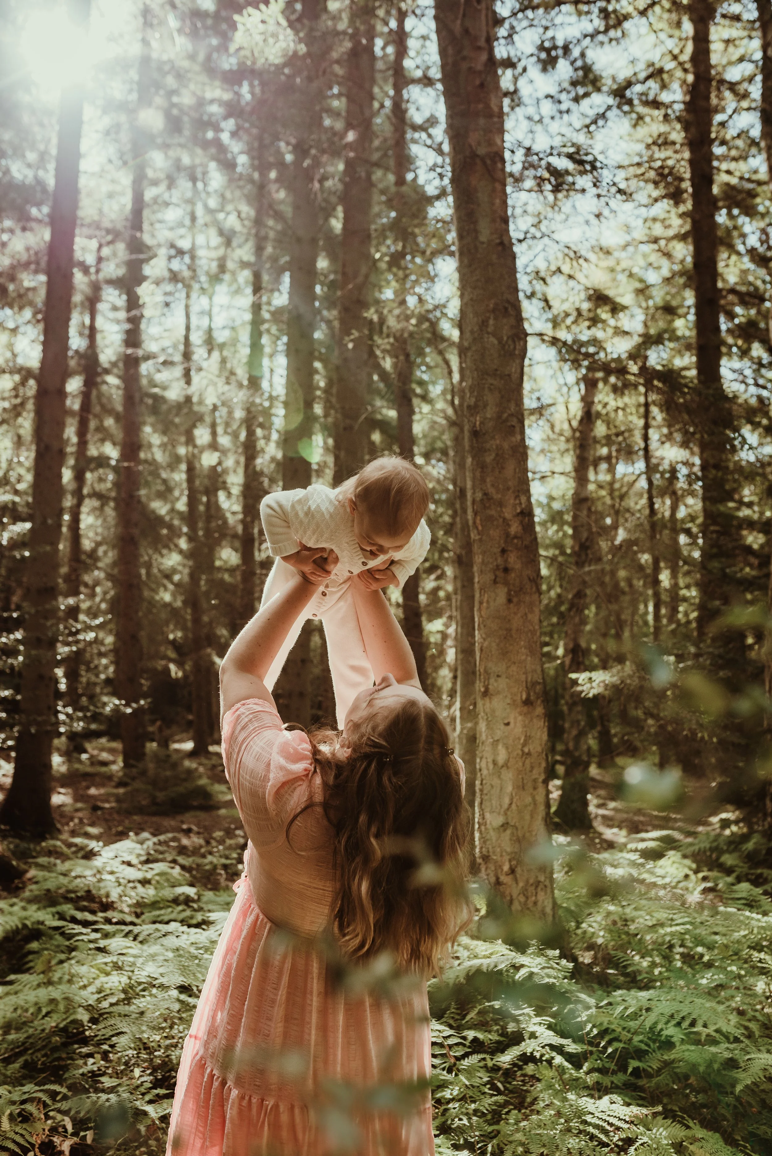 Mother lifting her baby joyfully in a Scottish forest in a pink dress during an outdoor nursing photography session with Marieke Captures Birth