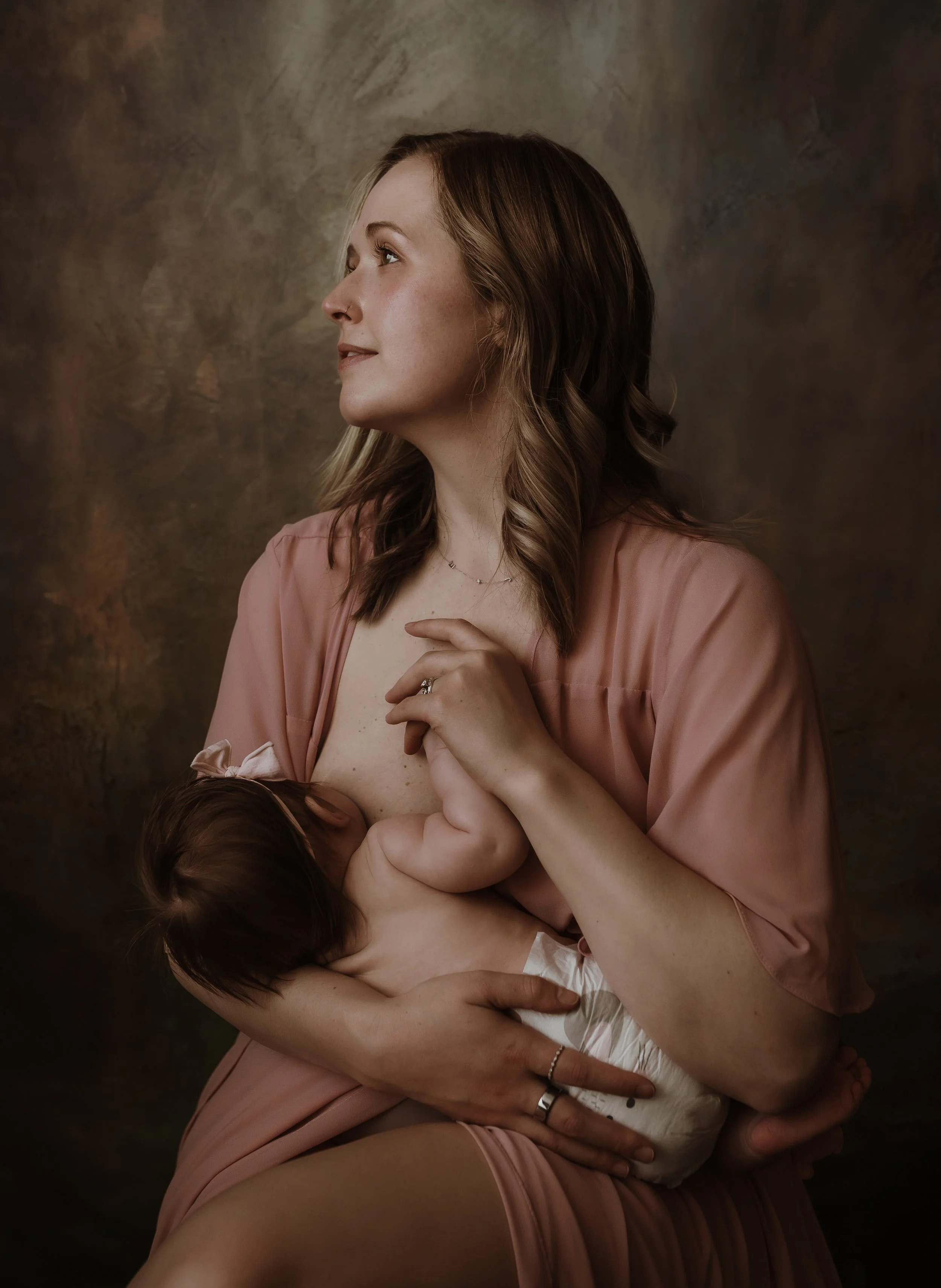 Studio portrait of a mother nursing her newborn while looking upward in a pink robe during a nursing photography session with Glasgow photographer Marieke Captures Birth