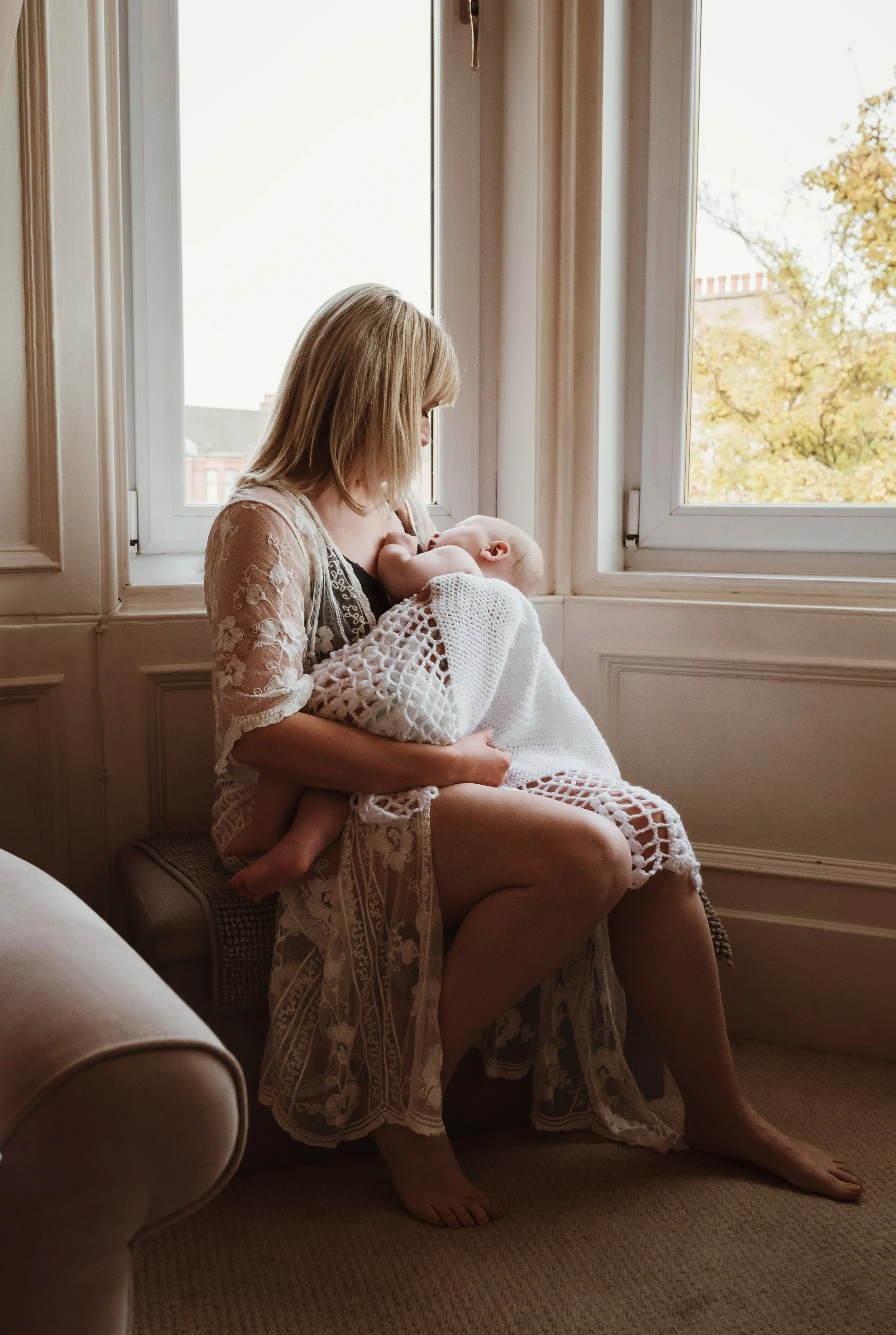 Mother nursing her baby by a sunlit window in a delicate lace top during a nursing photography session with Marieke Captures Birth Scotland