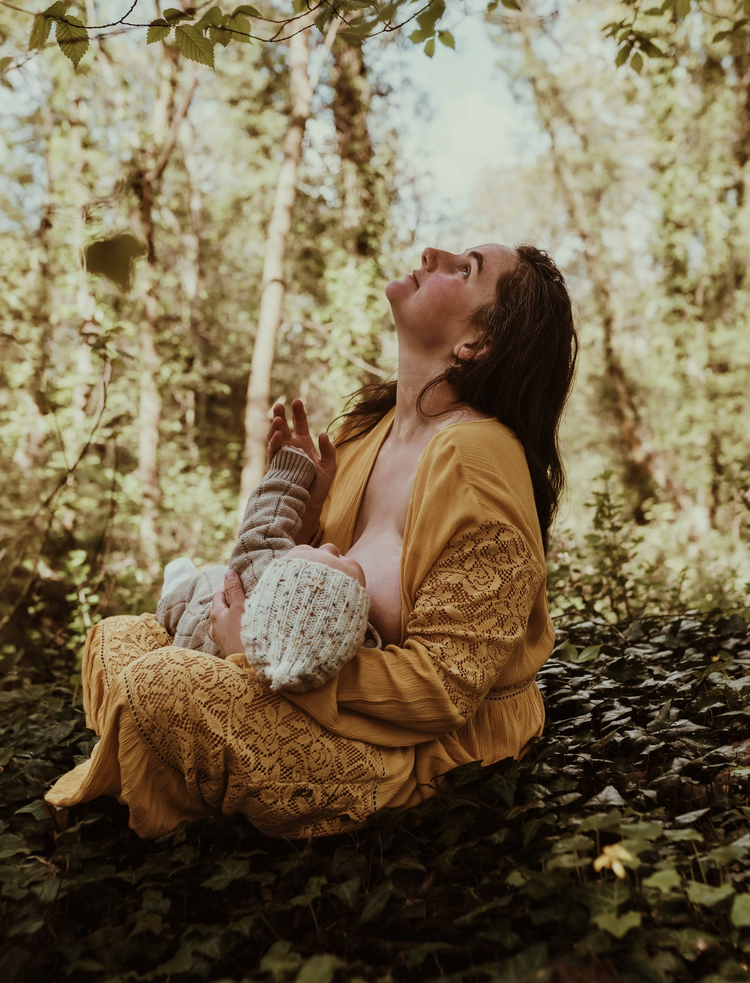 Mother nursing her baby while looking upward in a yellow dress during an outdoor nursing photography session in Scottish woodland with Marieke Captures Birth