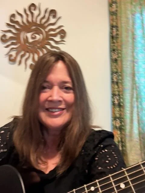 A woman with shoulder-length brown hair smiling, holding a guitar, sitting indoors near floral patterned curtains, with a sun and eye design wall decoration in the background.