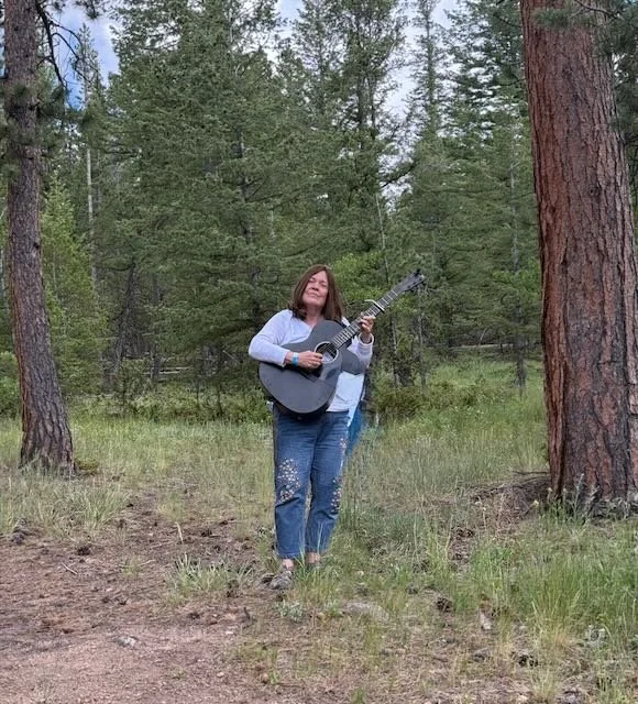 A woman standing in a forest, playing a black acoustic guitar, with a joyful expression.