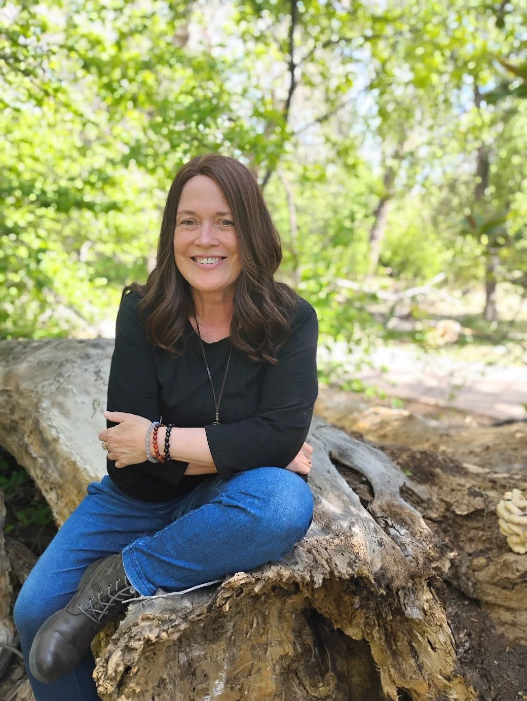 Anisha Zo, soul guide, evidential mediums, shamanic healing arts practitioner, reseacher and writing, studying high sensitivity and intuitive and spiritual giftedness. Smiling woman sitting on a fallen tree trunk in a lush green forest.