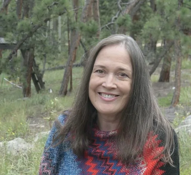 A woman with long gray hair smiling outdoors in a forested area, wearing a colorful sweater.